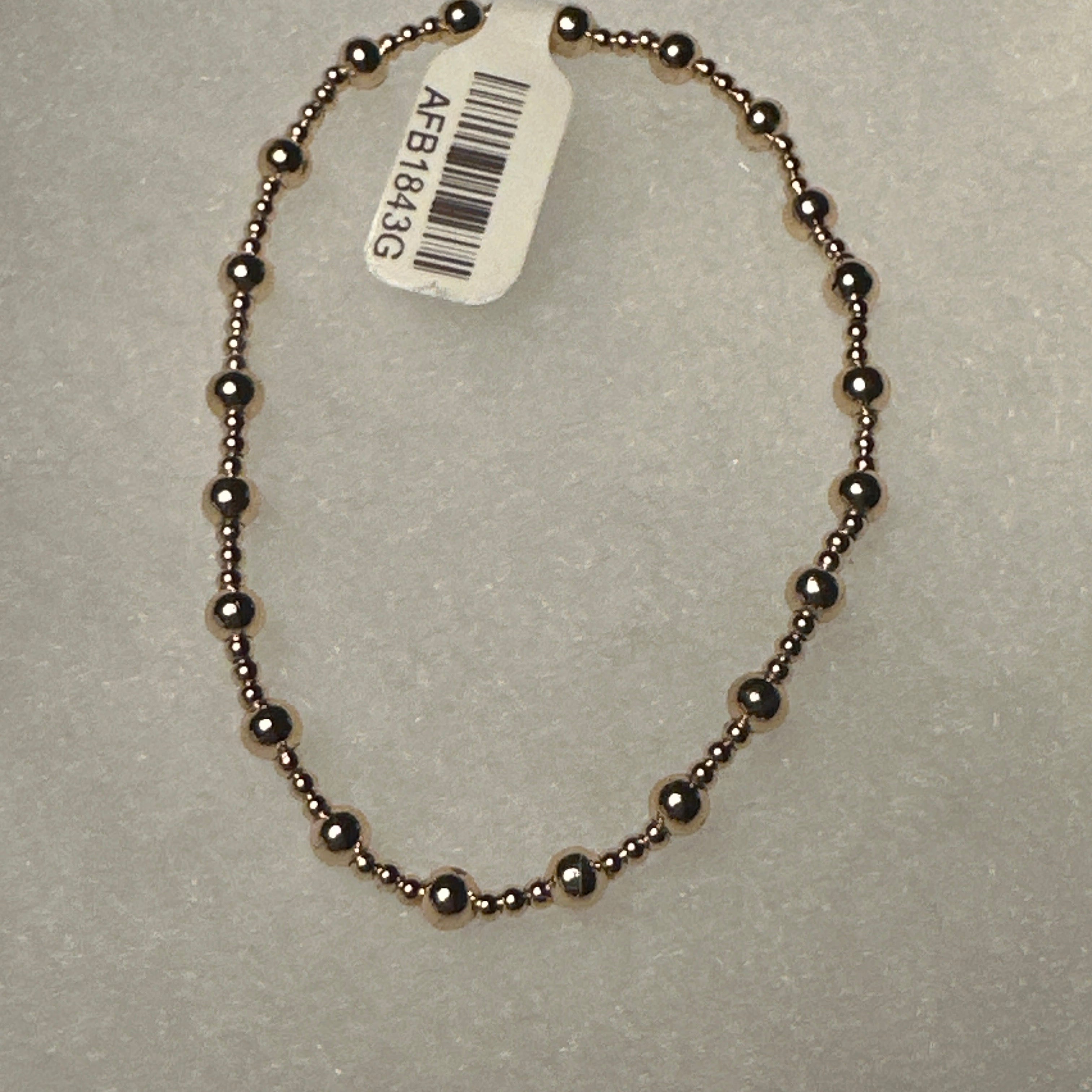 A beaded bracelet made of small metallic dark bronze beads, shown on a textured light gray background with a white product tag attached.