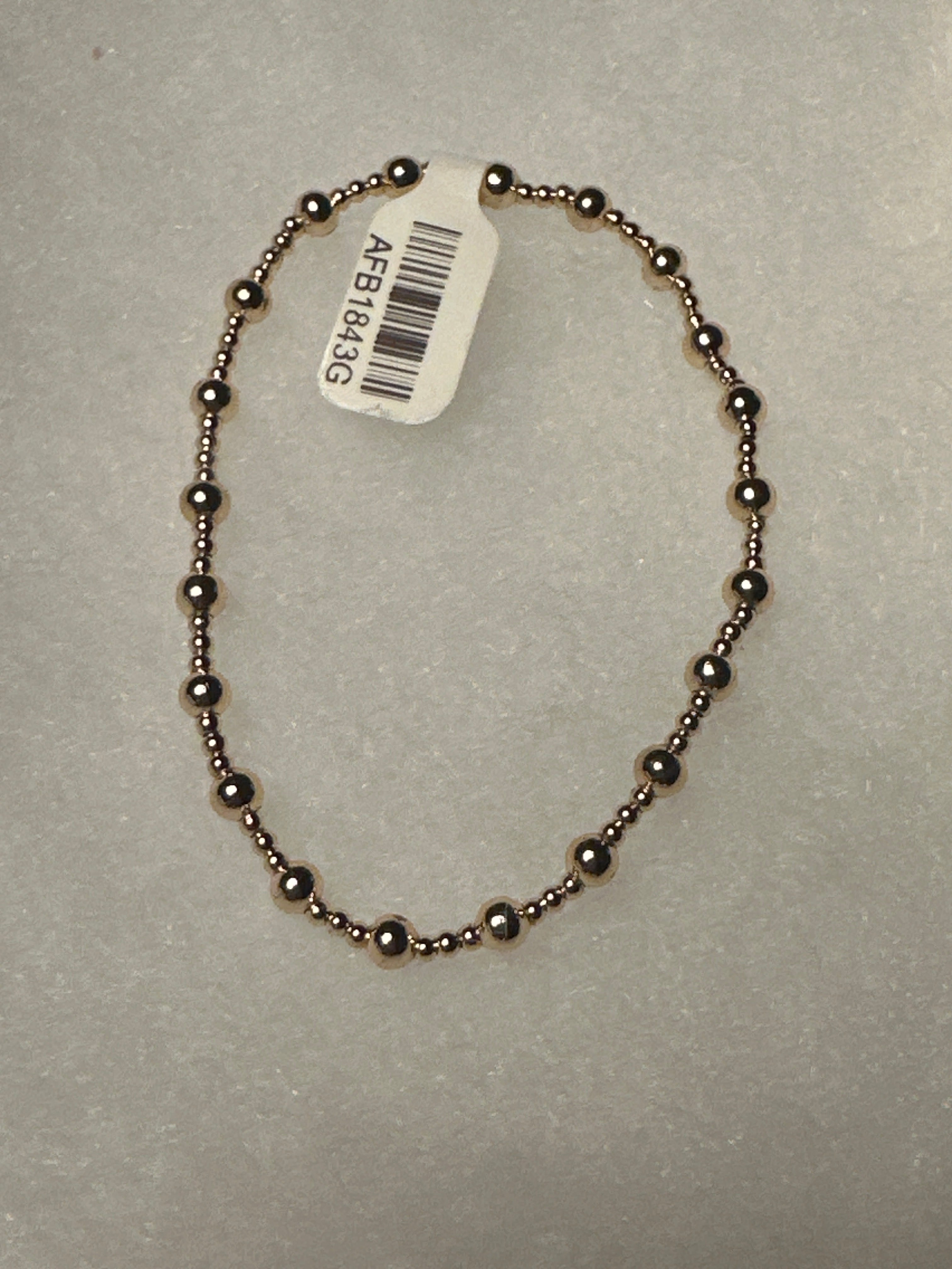 A beaded bracelet made of small metallic dark bronze beads, shown on a textured light gray background with a white product tag attached.