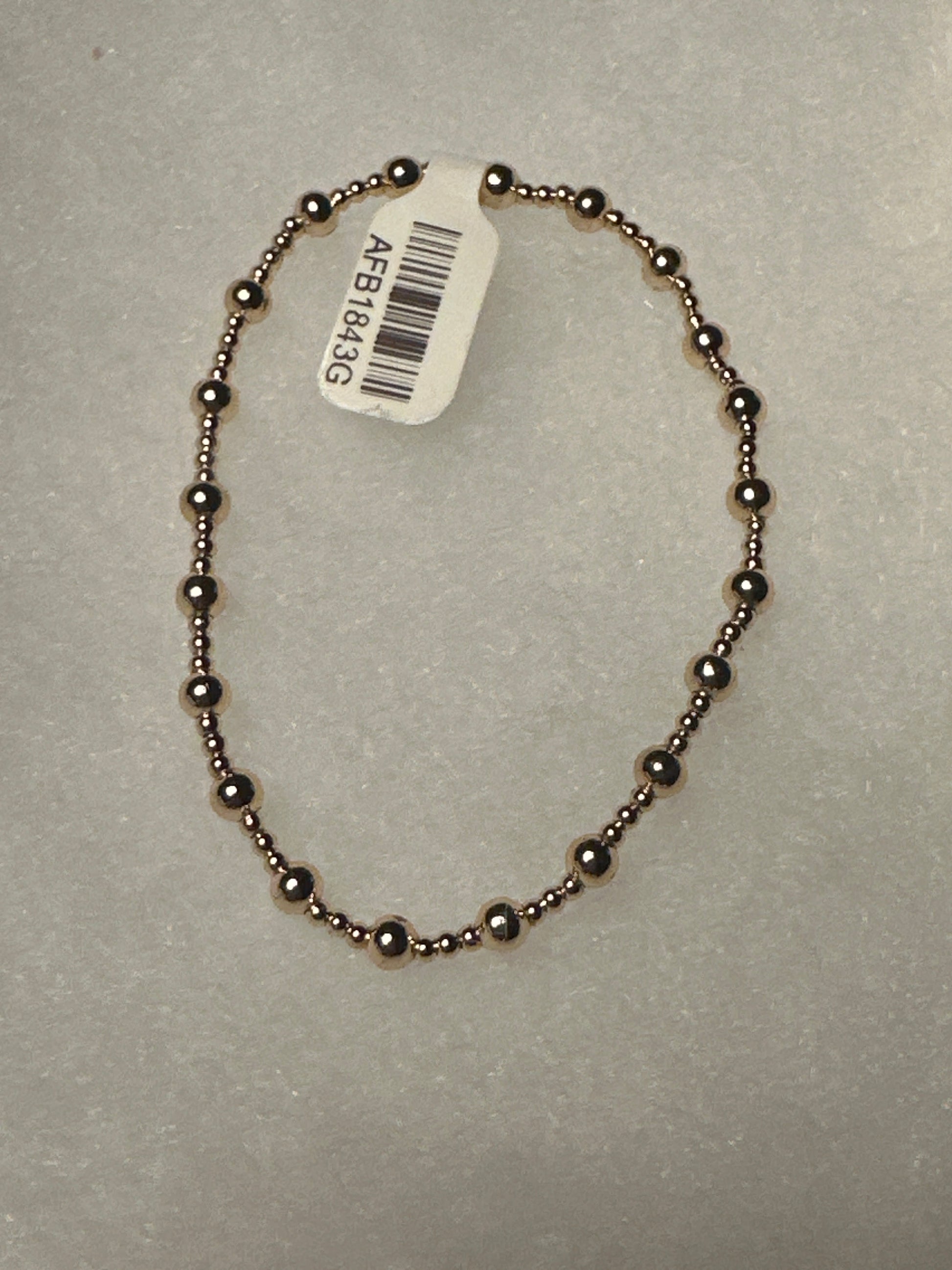 A beaded bracelet made of small metallic dark bronze beads, shown on a textured light gray background with a white product tag attached.