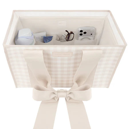 A square fabric storage organizer bin with a beige and white gingham pattern, featuring wide cream-colored shoulder straps tied into a large bow at the front. The open top of the bin is shown holding various small personal items like a phone, glasses, and a small bottle.