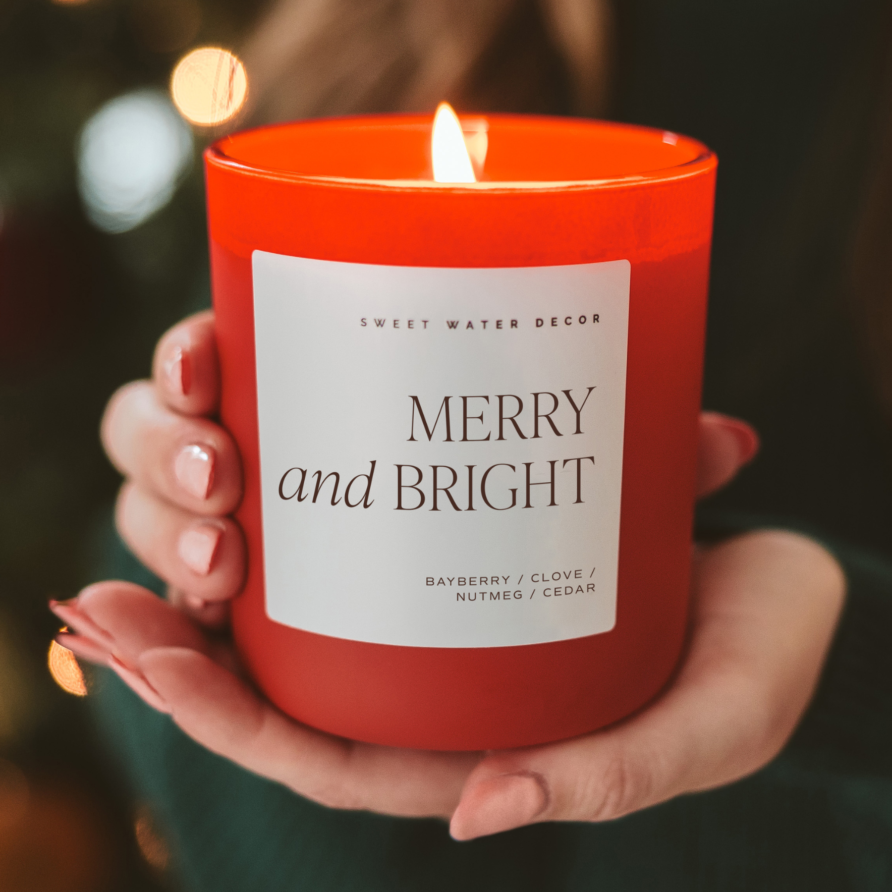 Red candle labeled 'Merry and Bright' held by a hand with a blurred festive background