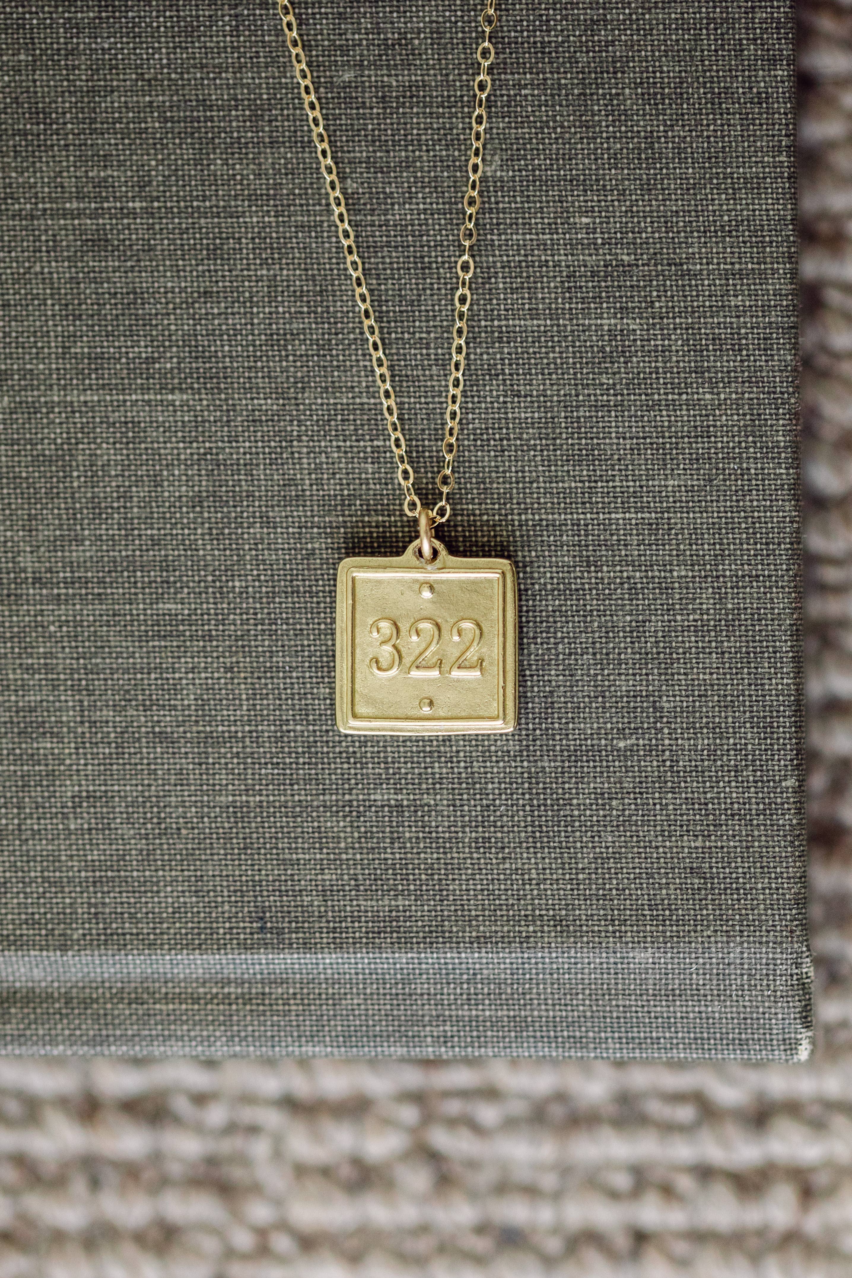 Gold necklace with a square pendant displaying the number '322' on a textured surface.