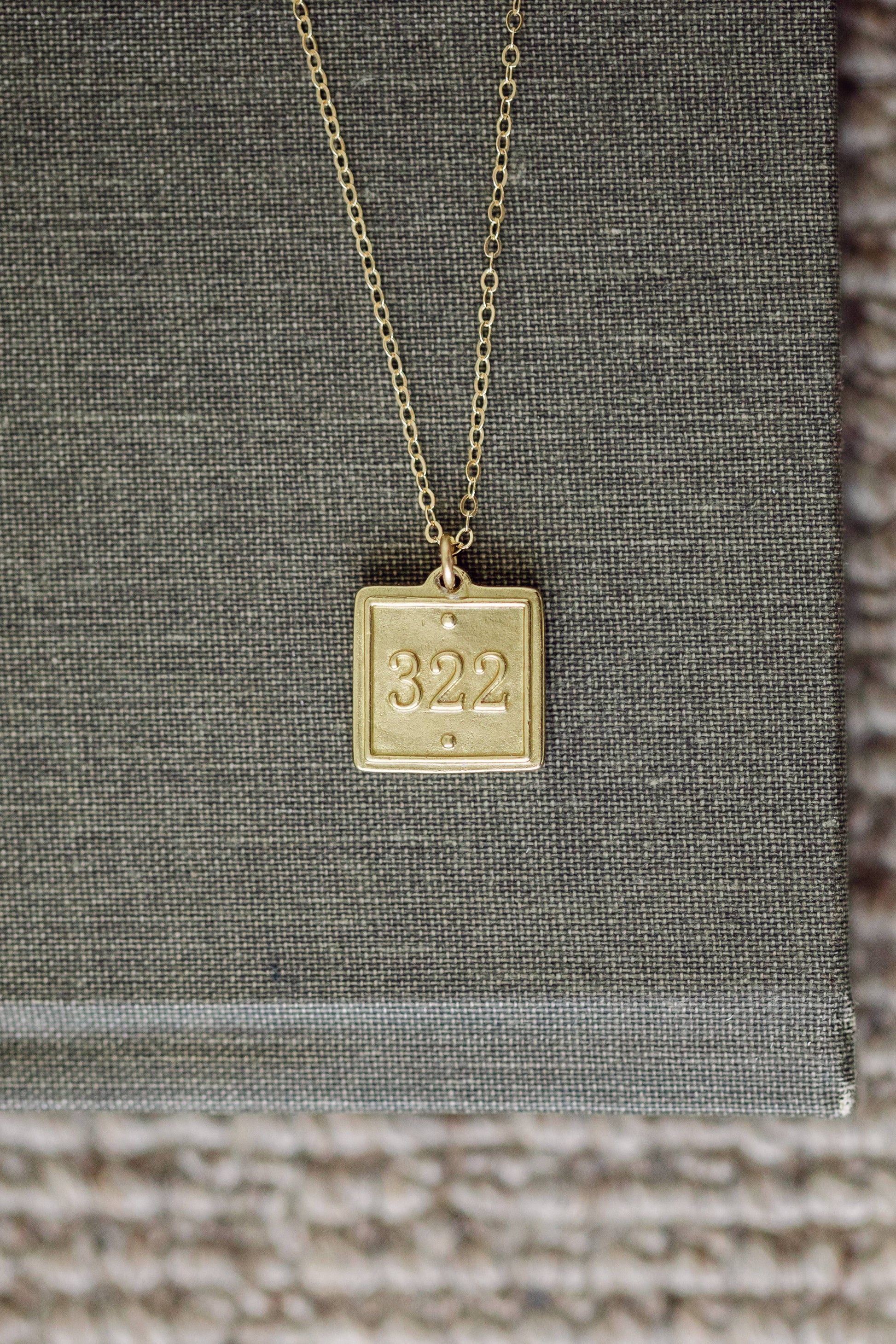 Gold necklace with a square pendant displaying the number '322' on a textured surface.