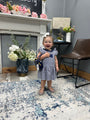 A toddler girl wearing a blue and white striped dress and holding a blue bag, standing in a decorated room with a fireplace, chair, and patterned rug.