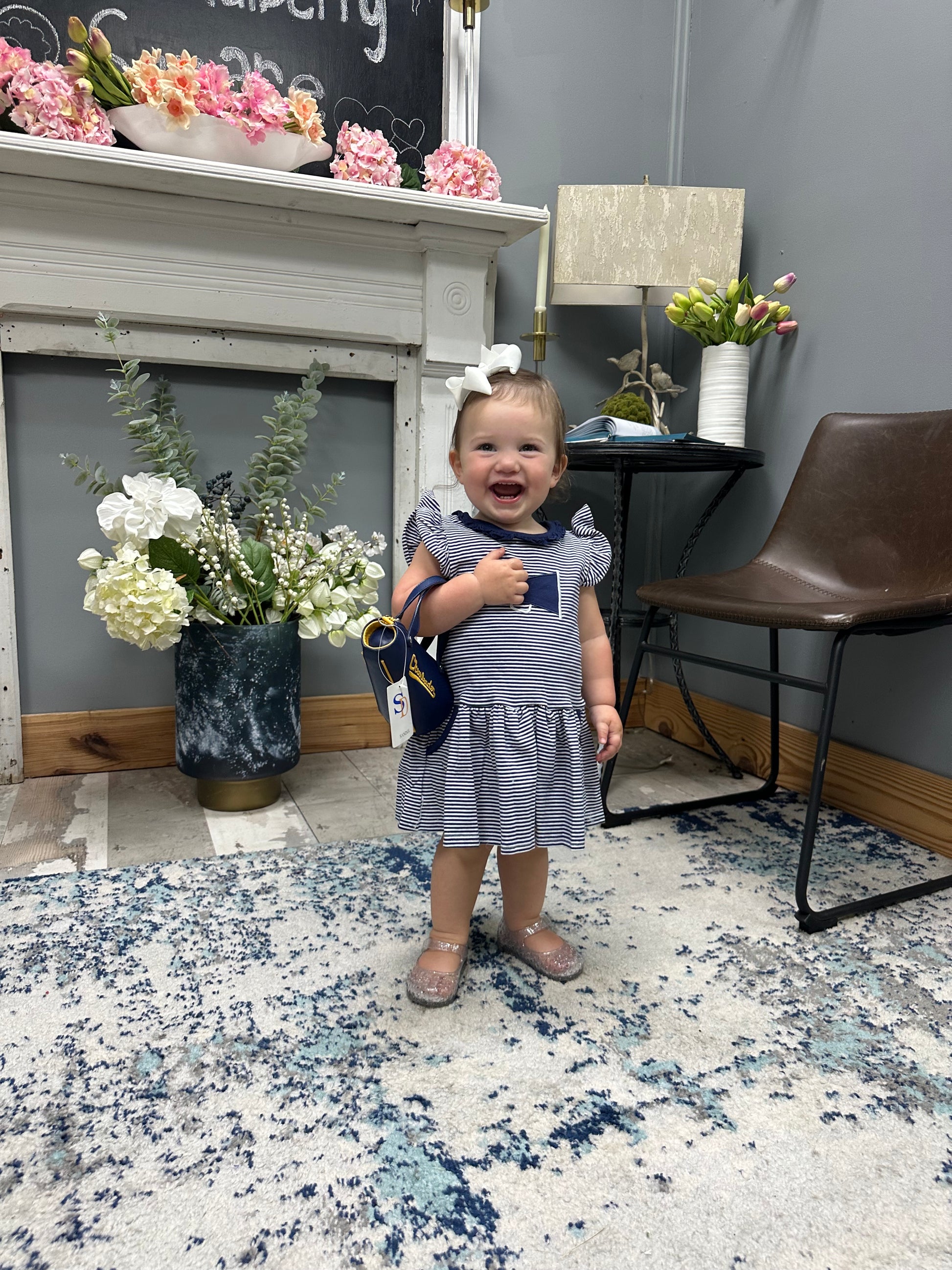 A toddler girl wearing a blue and white striped dress and holding a blue bag, standing in a decorated room with a fireplace, chair, and patterned rug.