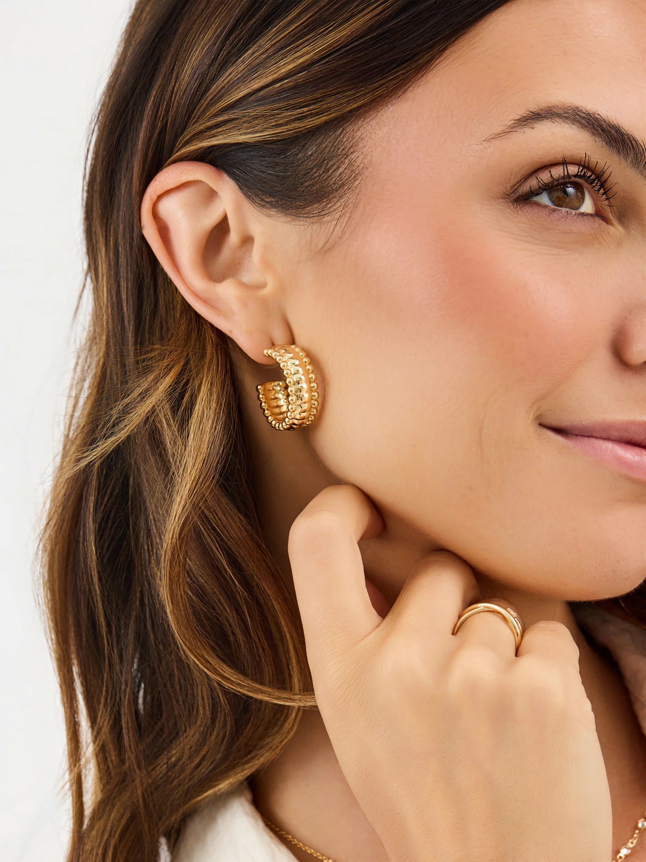 Close-up of a woman wearing gold hoop earrings and a ring, with a plain background.