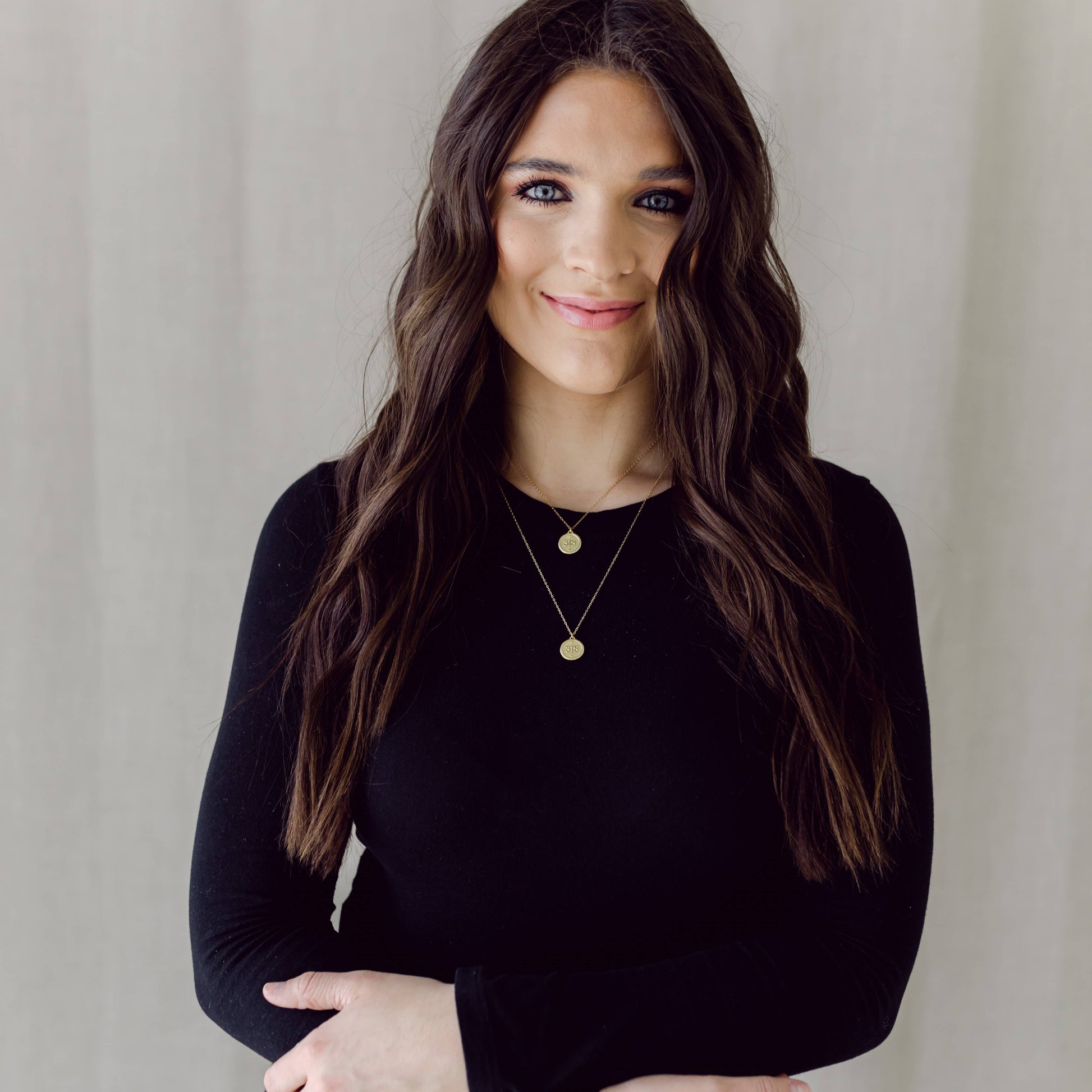 Woman wearing a black long-sleeve top with gold jewelry against a neutral background