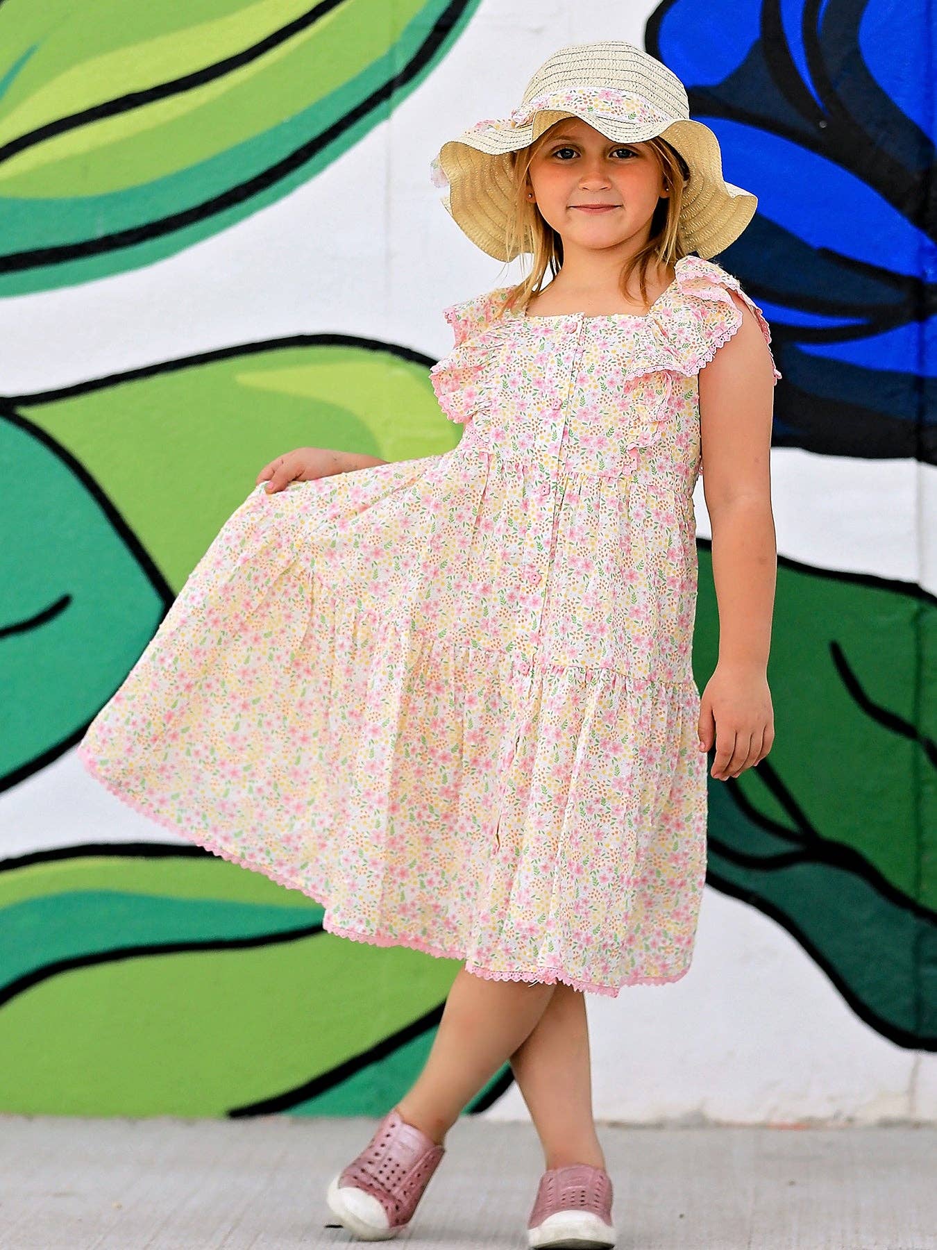 Young girl in a floral dress and sun hat standing in front of a colorful mural.