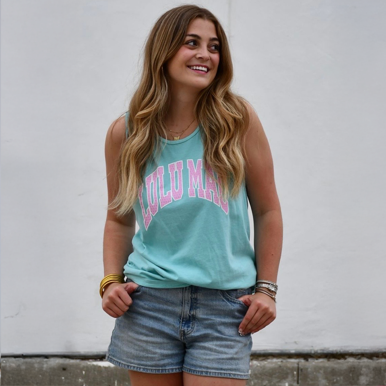 Woman wearing a turquoise tank top with pink text and denim shorts against a white wall.