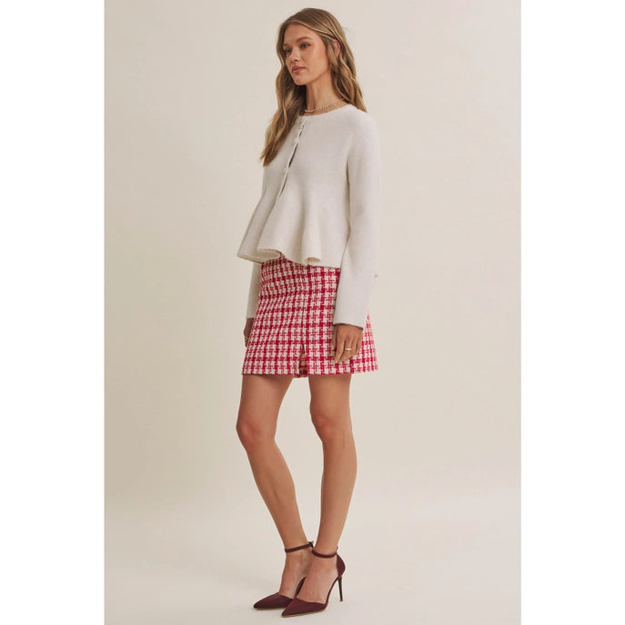 A woman modeling a light beige cropped cardigan and a red and white gingham mini skirt, paired with burgundy high heels, against a solid beige studio background.