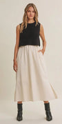 A woman modeling a sleeveless black ribbed knit crop top and a high-waisted, pleated off-white midi skirt with side pockets, paired with black ankle boots, against a plain beige studio background.