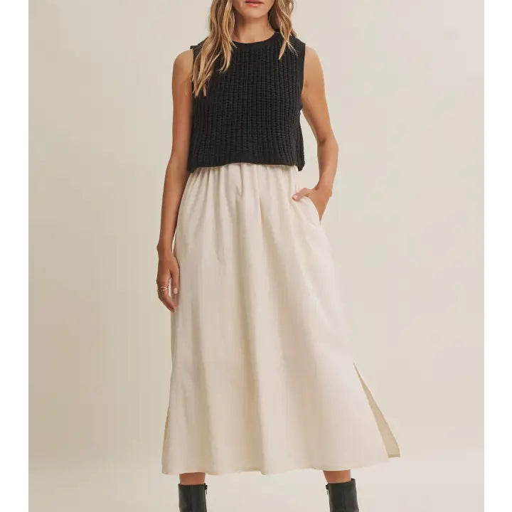 A woman modeling a sleeveless black ribbed knit crop top and a high-waisted, pleated off-white midi skirt with side pockets, paired with black ankle boots, against a plain beige studio background.