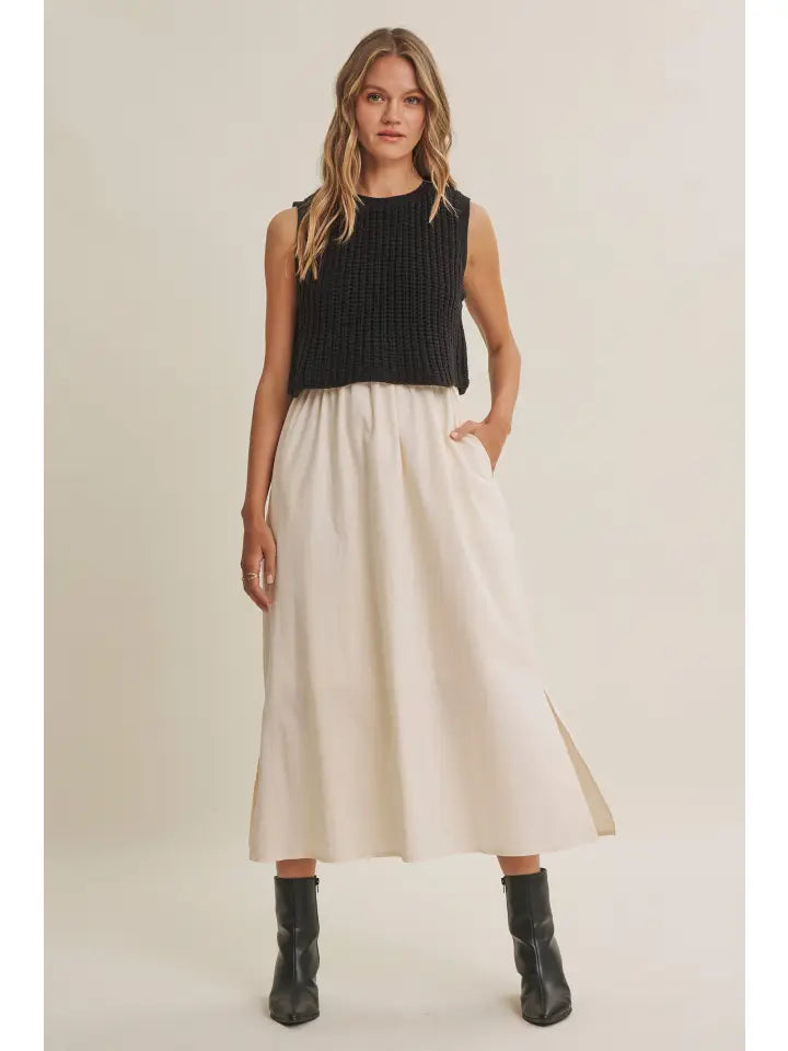 A woman modeling a sleeveless black ribbed knit crop top and a high-waisted, pleated off-white midi skirt with side pockets, paired with black ankle boots, against a plain beige studio background.