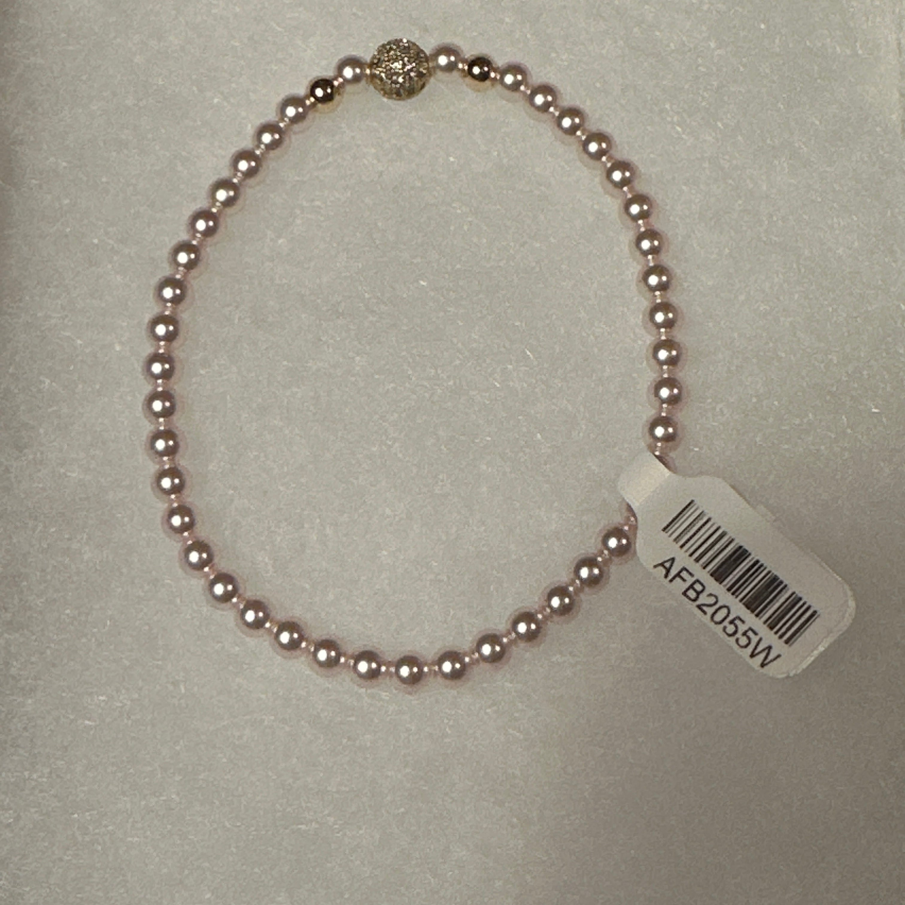 A single-strand pink pearl bracelet with a gold-toned decorative accent bead and a white product tag attached, displayed on a white textured surface.