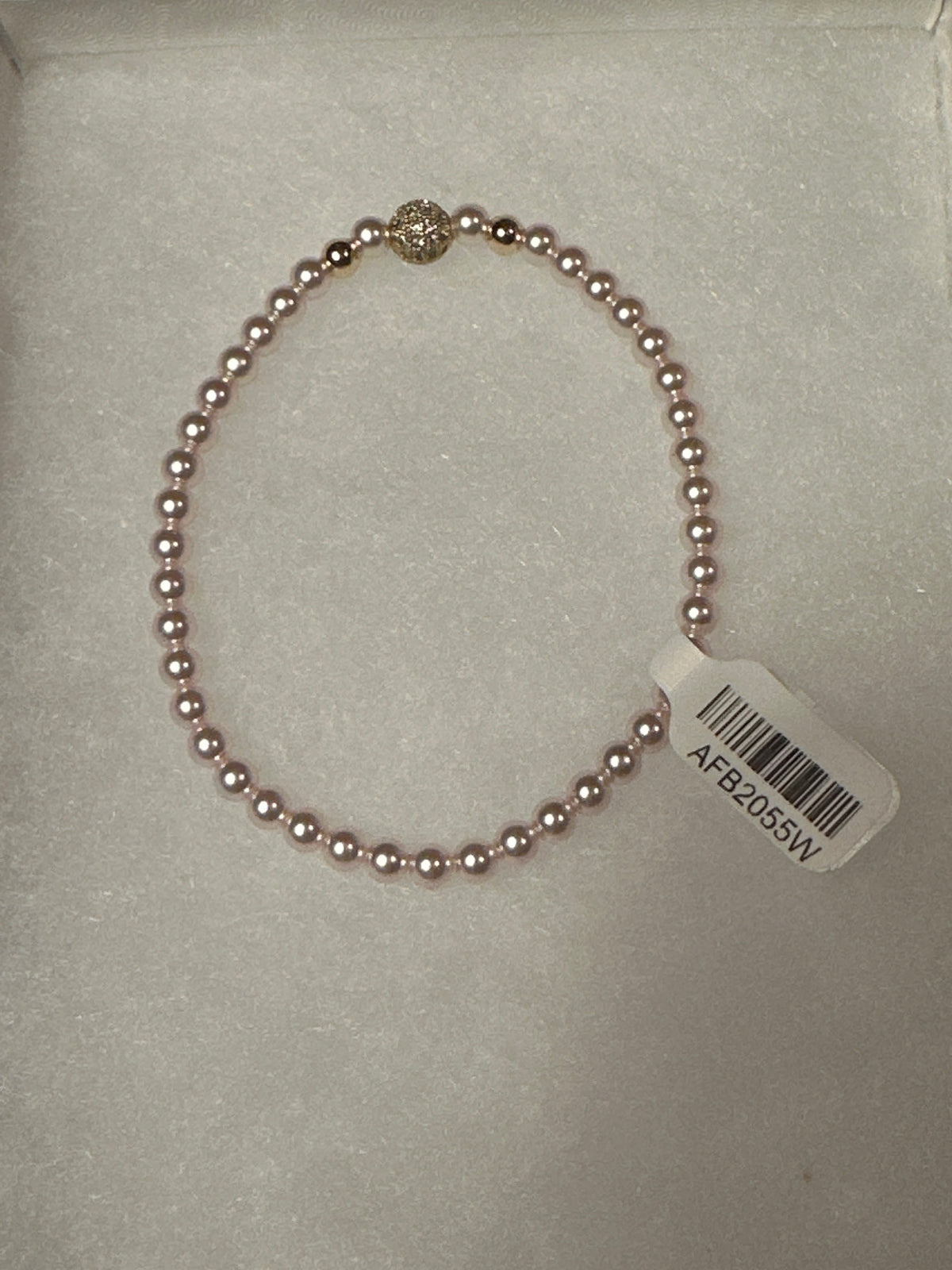 A single-strand pink pearl bracelet with a gold-toned decorative accent bead and a white product tag attached, displayed on a white textured surface.
