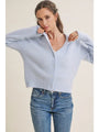 A woman wearing a light blue cropped button-up cardigan sweater and blue jeans, posing against a solid beige background.