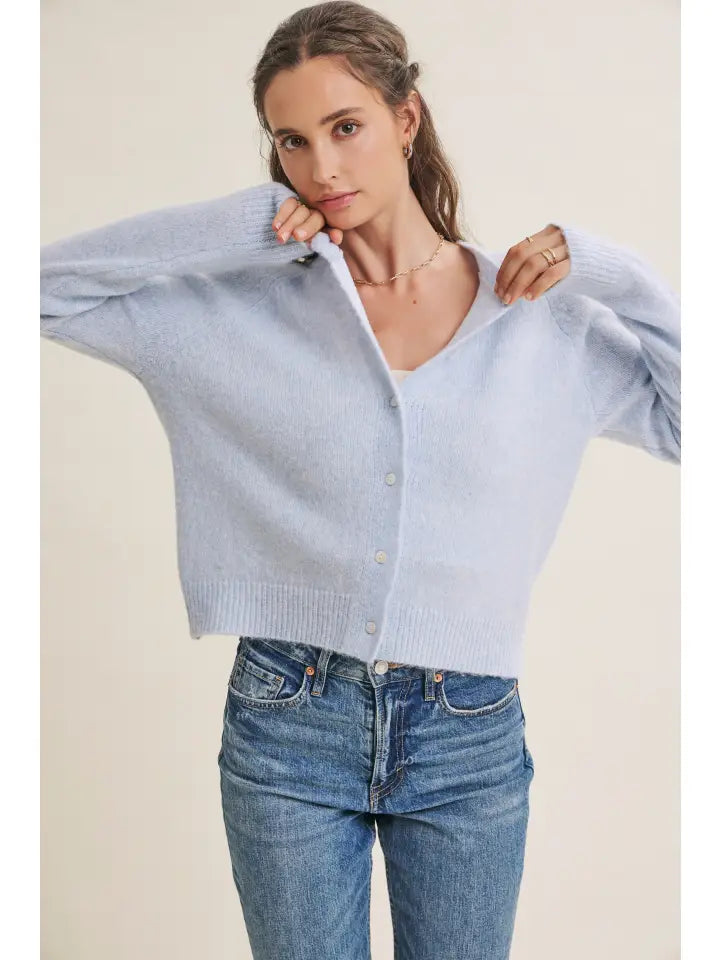 A woman wearing a light blue cropped button-up cardigan sweater and blue jeans, posing against a solid beige background.