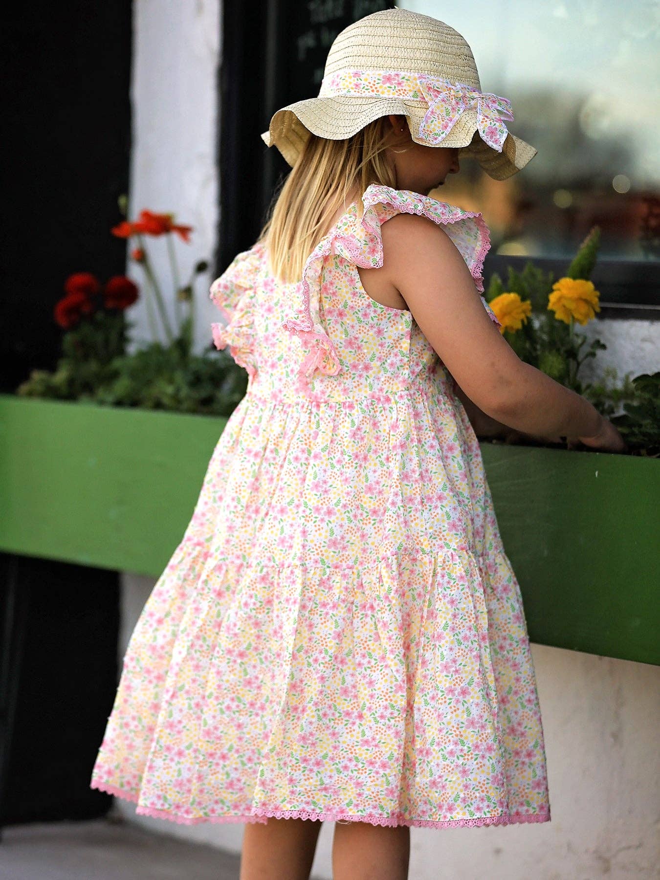 Young girl in a floral dress and sun hat standing near a planter with flowers.
