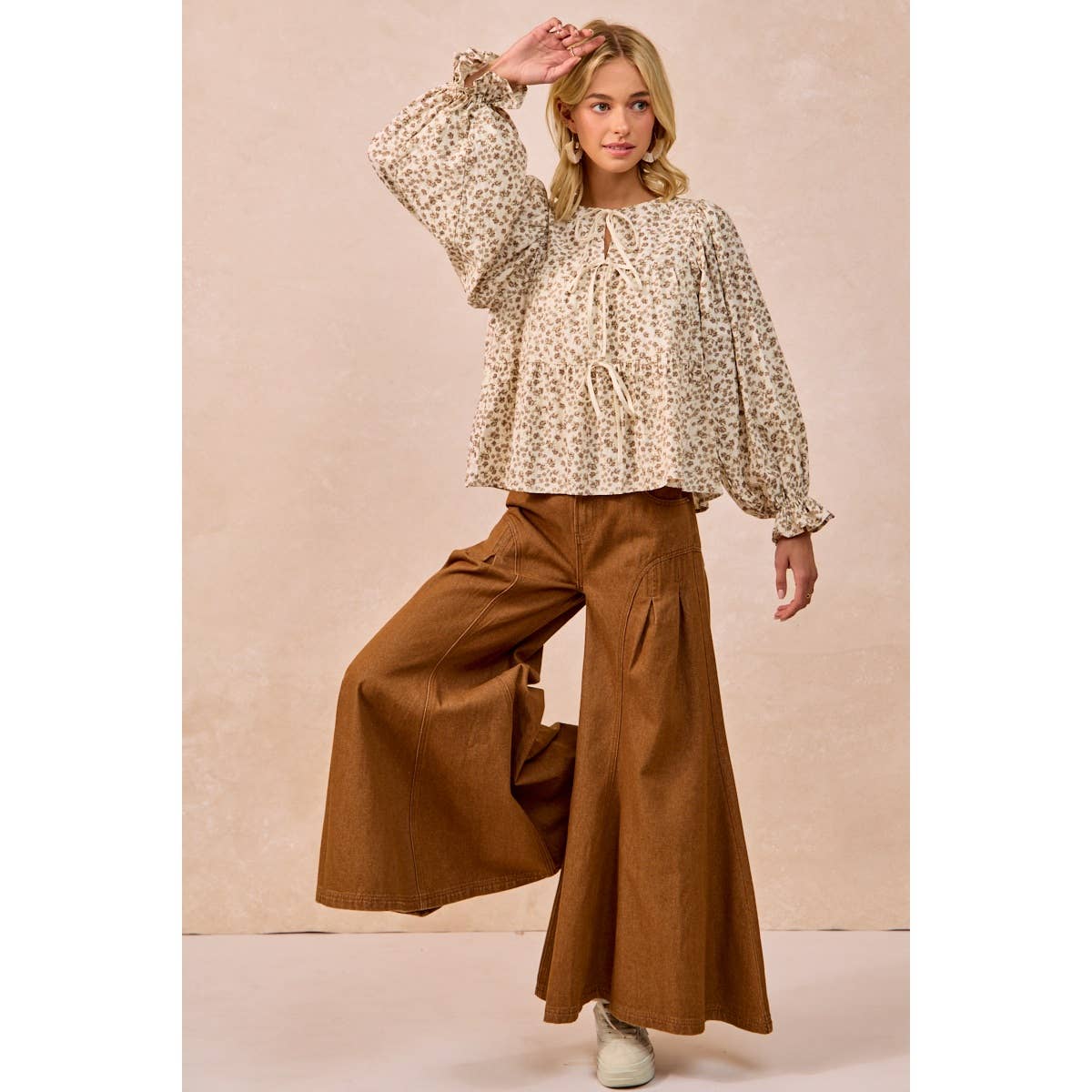 A woman modeling a beige and brown floral print long-sleeve blouse with a drawstring tie at the collar, paired with wide-leg brown pants, against a textured beige studio backdrop.