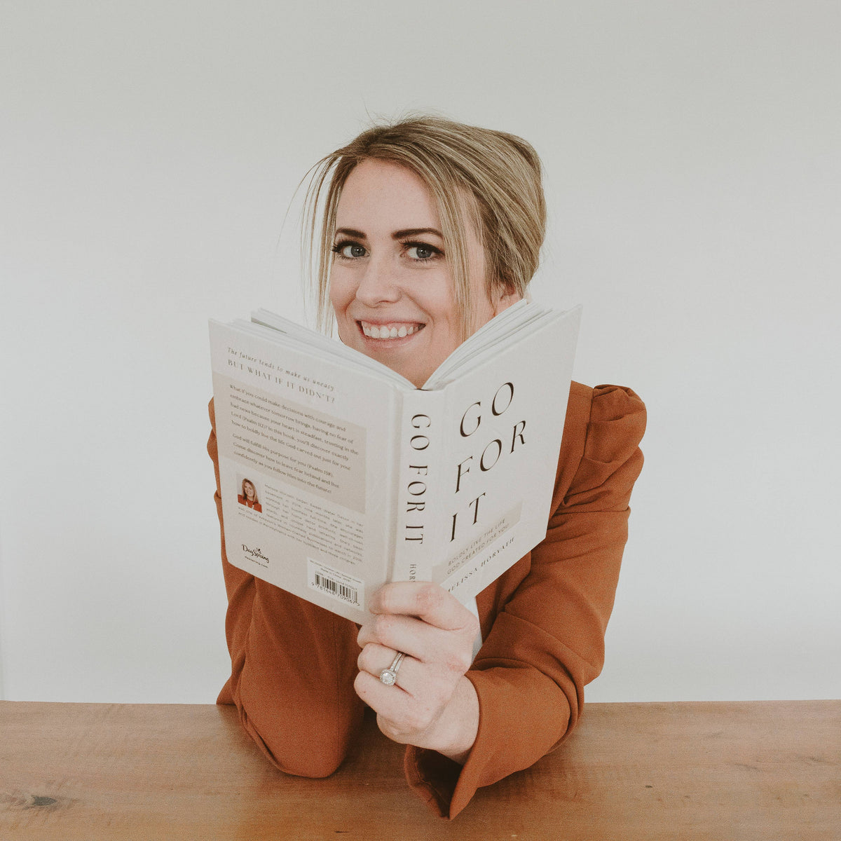 Woman holding a devotional book with both hands, smiling in rust-orange sleeves