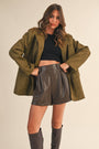Woman wearing a green coat, black top, and leather shorts on a beige background