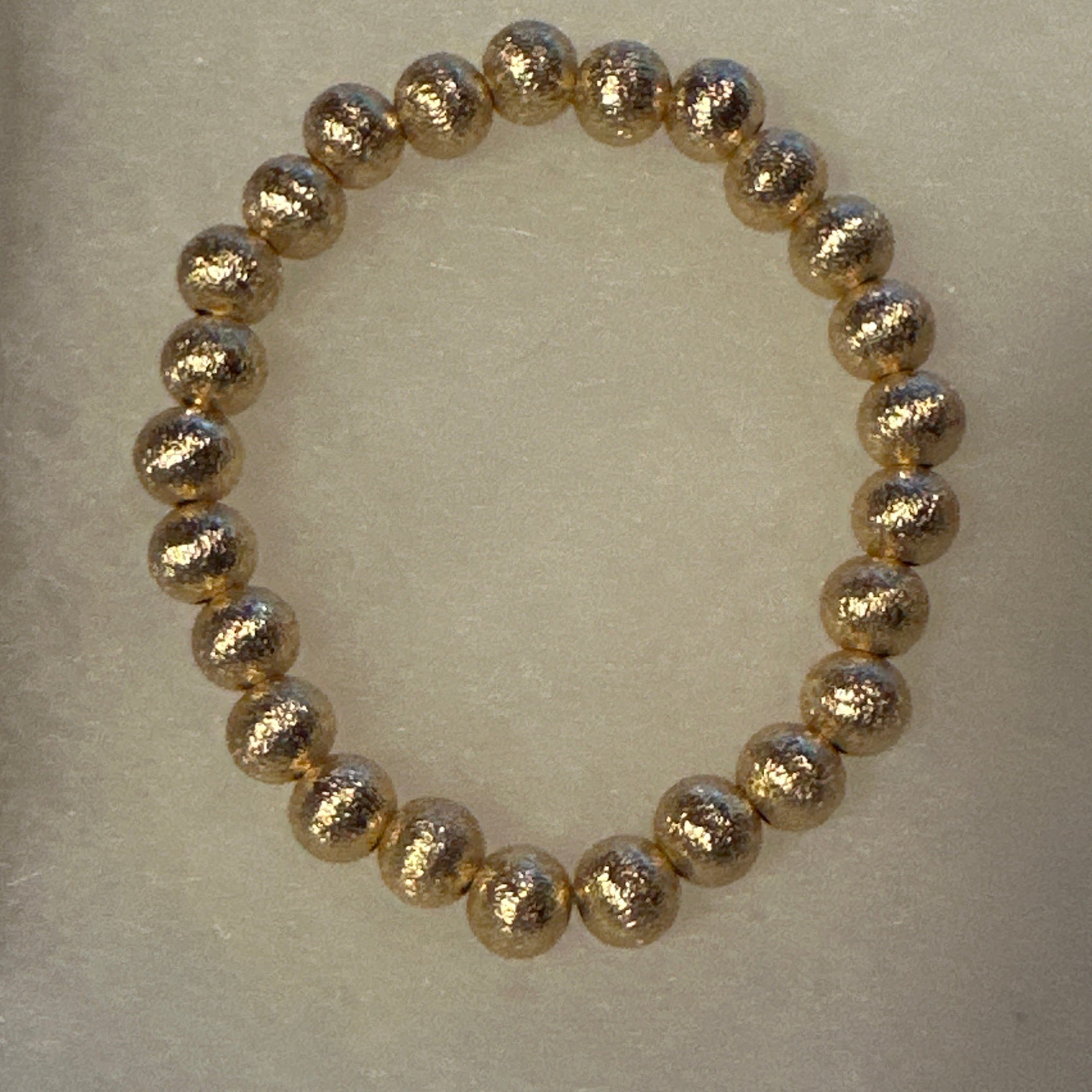 A beaded stretch bracelet made of metallic gold textured round beads, displayed on a neutral light beige background.