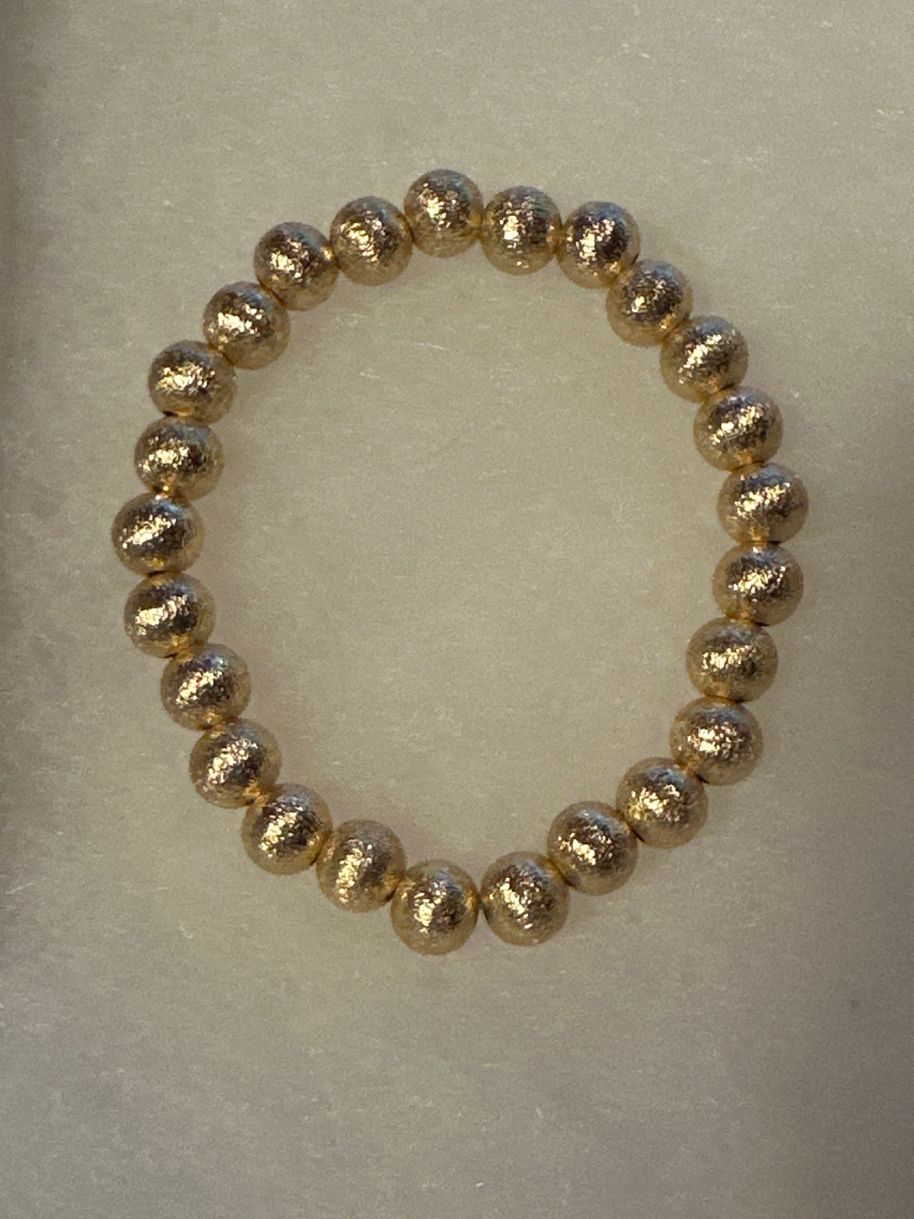 A beaded stretch bracelet made of metallic gold textured round beads, displayed on a neutral light beige background.