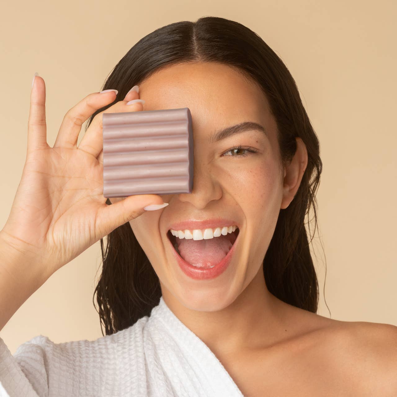 Woman holding a bar of soap in front of her eye on a beige background