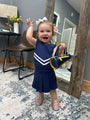 A toddler girl wearing a navy blue dress with white chevron stripes and holding a matching navy and yellow handbag, standing in a room with a mirror and wooden furniture.