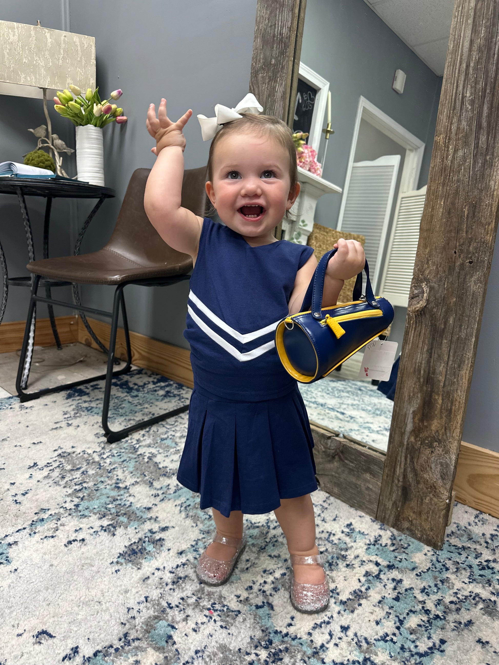 A toddler girl wearing a navy blue dress with white chevron stripes and holding a matching navy and yellow handbag, standing in a room with a mirror and wooden furniture.