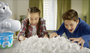 Two children playing with a large pile of white foam balls in a room with a toy and bucket visible.