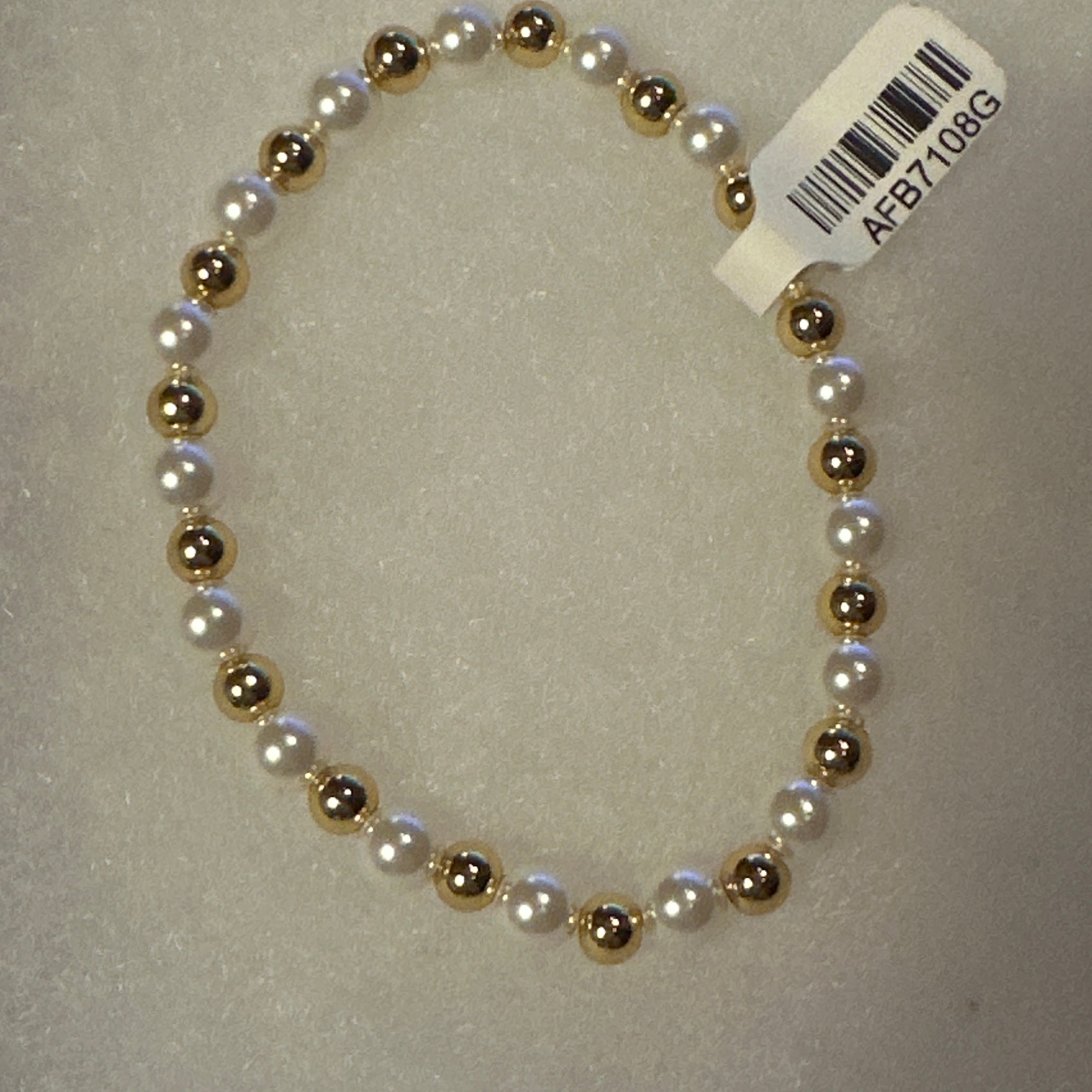 A beaded stretch bracelet featuring alternating white faux pearls and metallic gold beads, with a white barcode tag attached, shown on a textured light gray background.