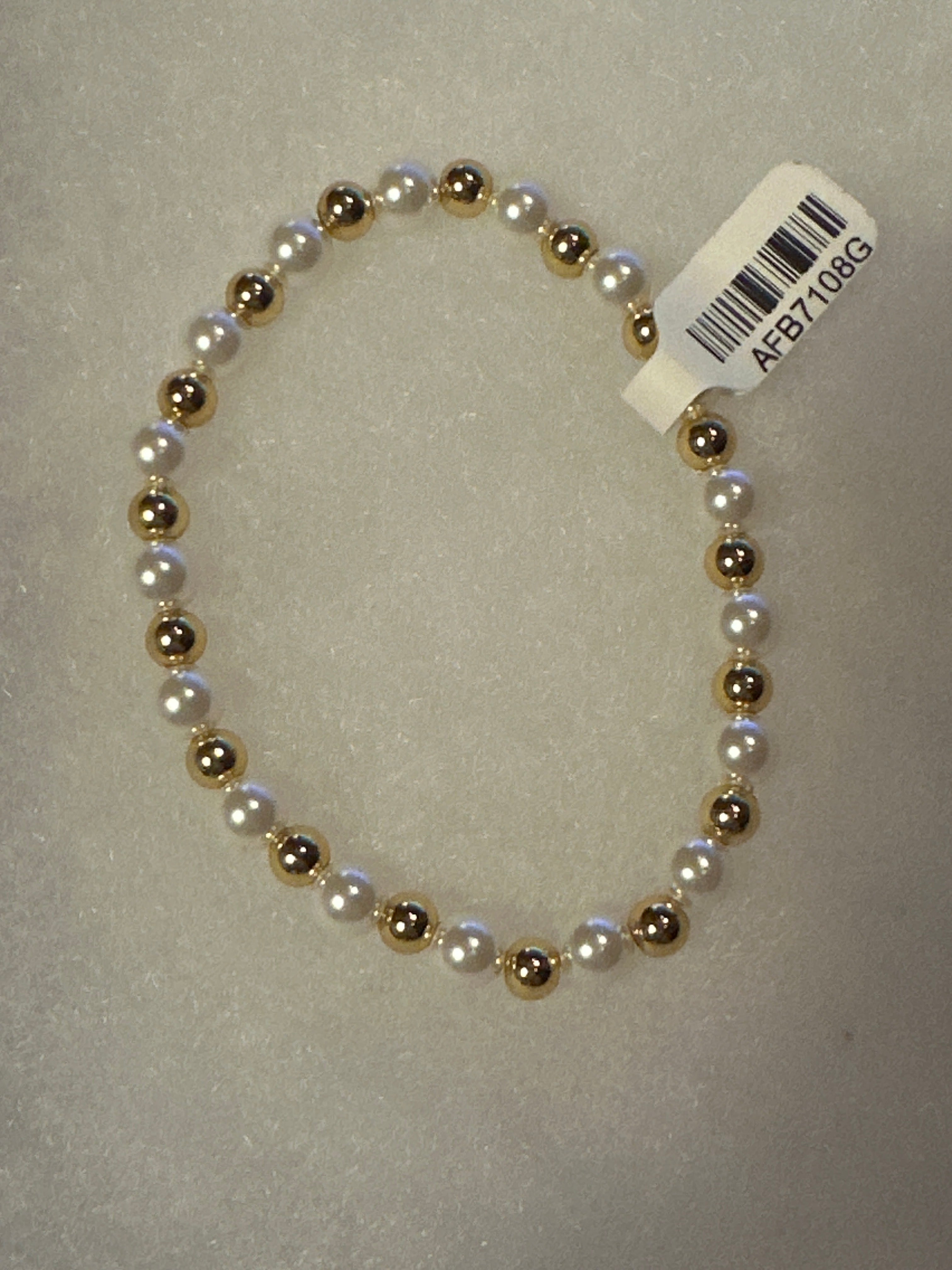 A beaded stretch bracelet featuring alternating white faux pearls and metallic gold beads, with a white barcode tag attached, shown on a textured light gray background.