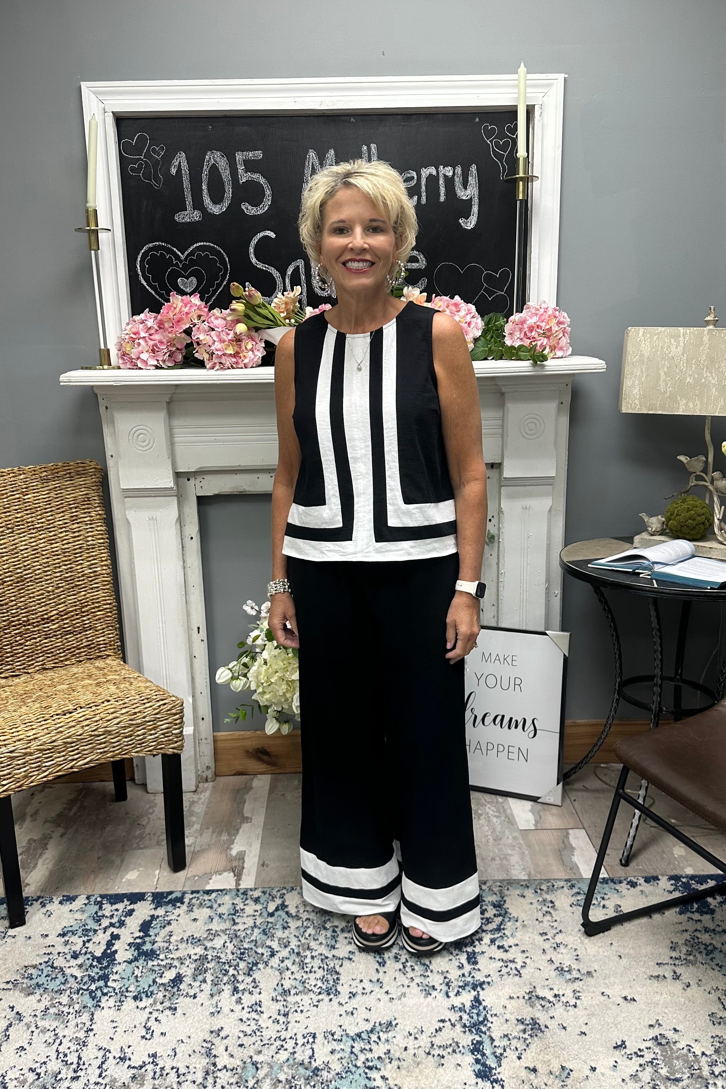 Woman in a black and white outfit standing in a room with a fireplace and decorative items.