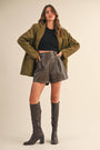 Woman wearing an olive green coat, black top, gray leather shorts, and black knee-high boots on a beige background