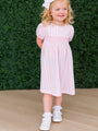 Young girl in a pink striped dress standing against a green hedge.