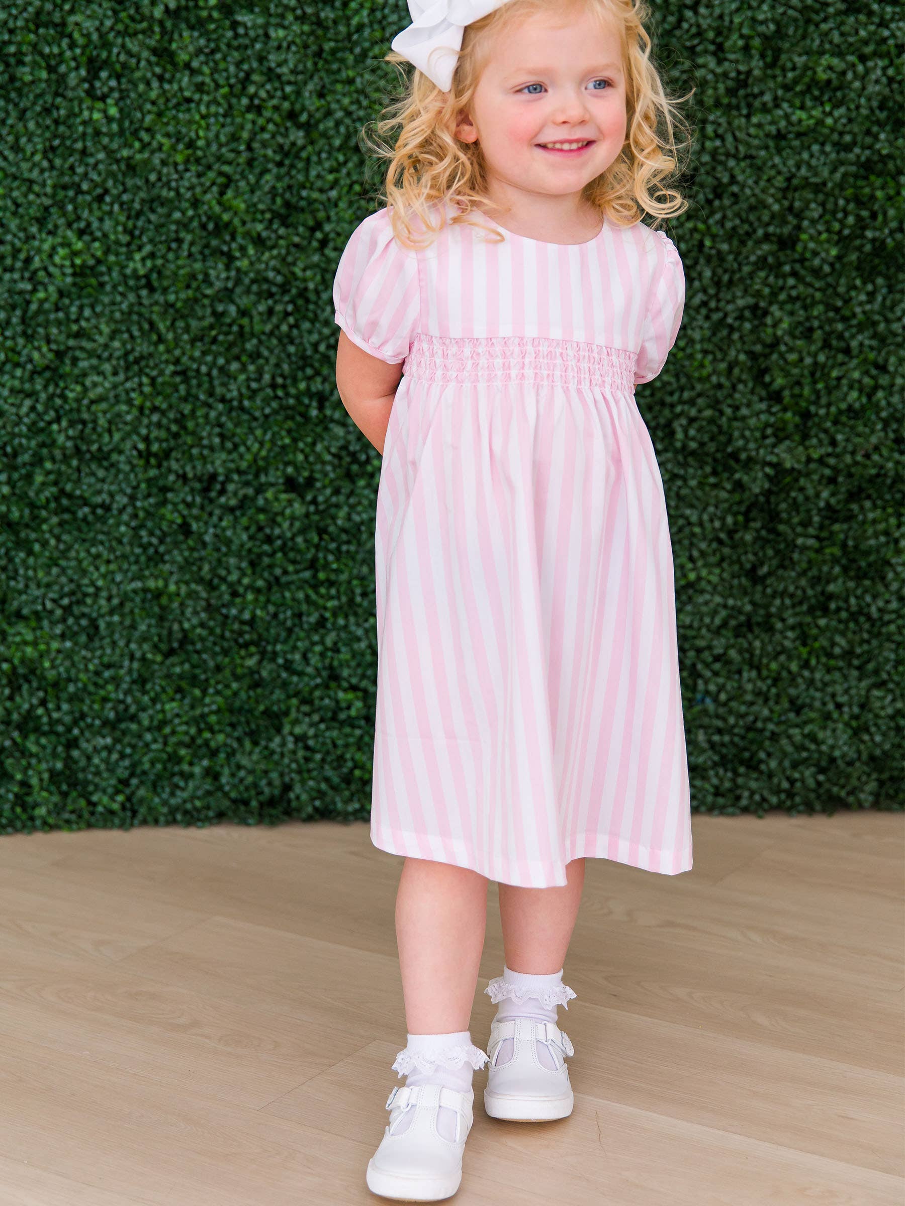 Young girl in a pink striped dress standing against a green hedge.