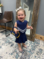 A toddler girl wearing a navy blue and white cheerleader-style dress and silver glittery shoes, holding a blue and yellow 'Charades' purse, standing in a room with a patterned rug and a wooden mirror.