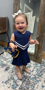 A toddler girl wearing a navy blue and white cheerleader-style dress and silver glittery shoes, holding a blue and yellow 'Charades' purse, standing in a room with a patterned rug and a wooden mirror.
