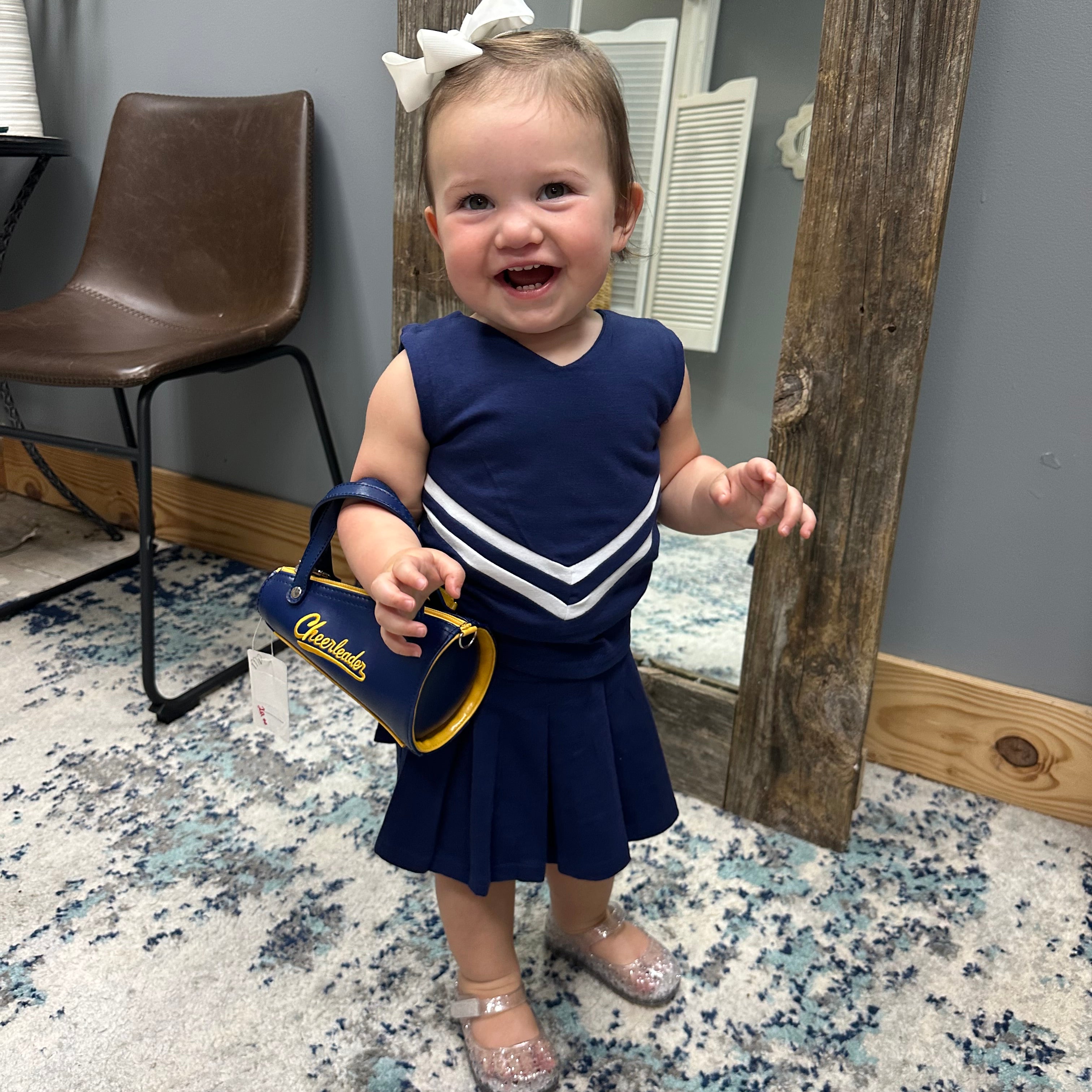A toddler girl wearing a navy blue and white cheerleader-style dress and silver glittery shoes, holding a blue and yellow 'Charades' purse, standing in a room with a patterned rug and a wooden mirror.