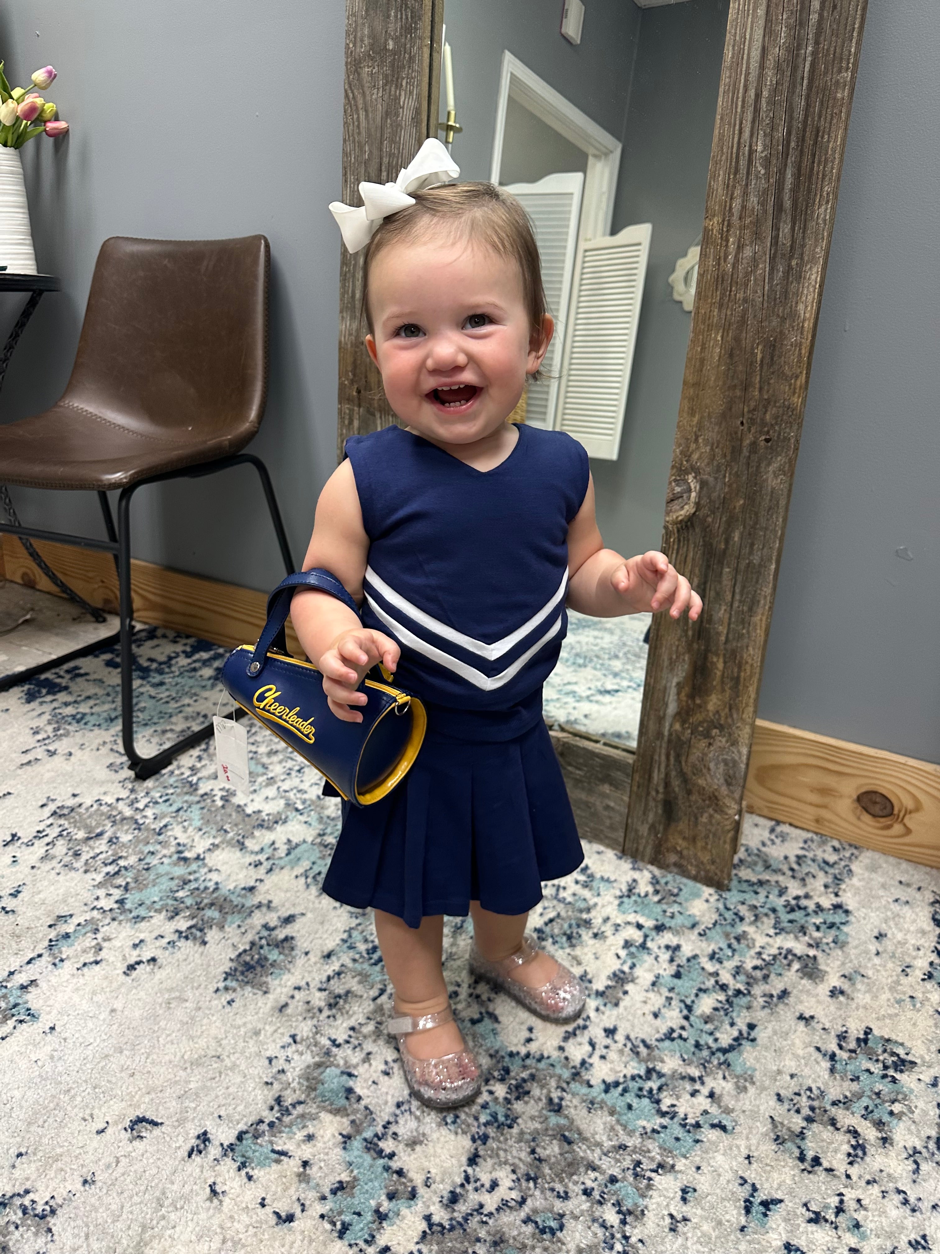 A toddler girl wearing a navy blue and white cheerleader-style dress and silver glittery shoes, holding a blue and yellow 'Charades' purse, standing in a room with a patterned rug and a wooden mirror.