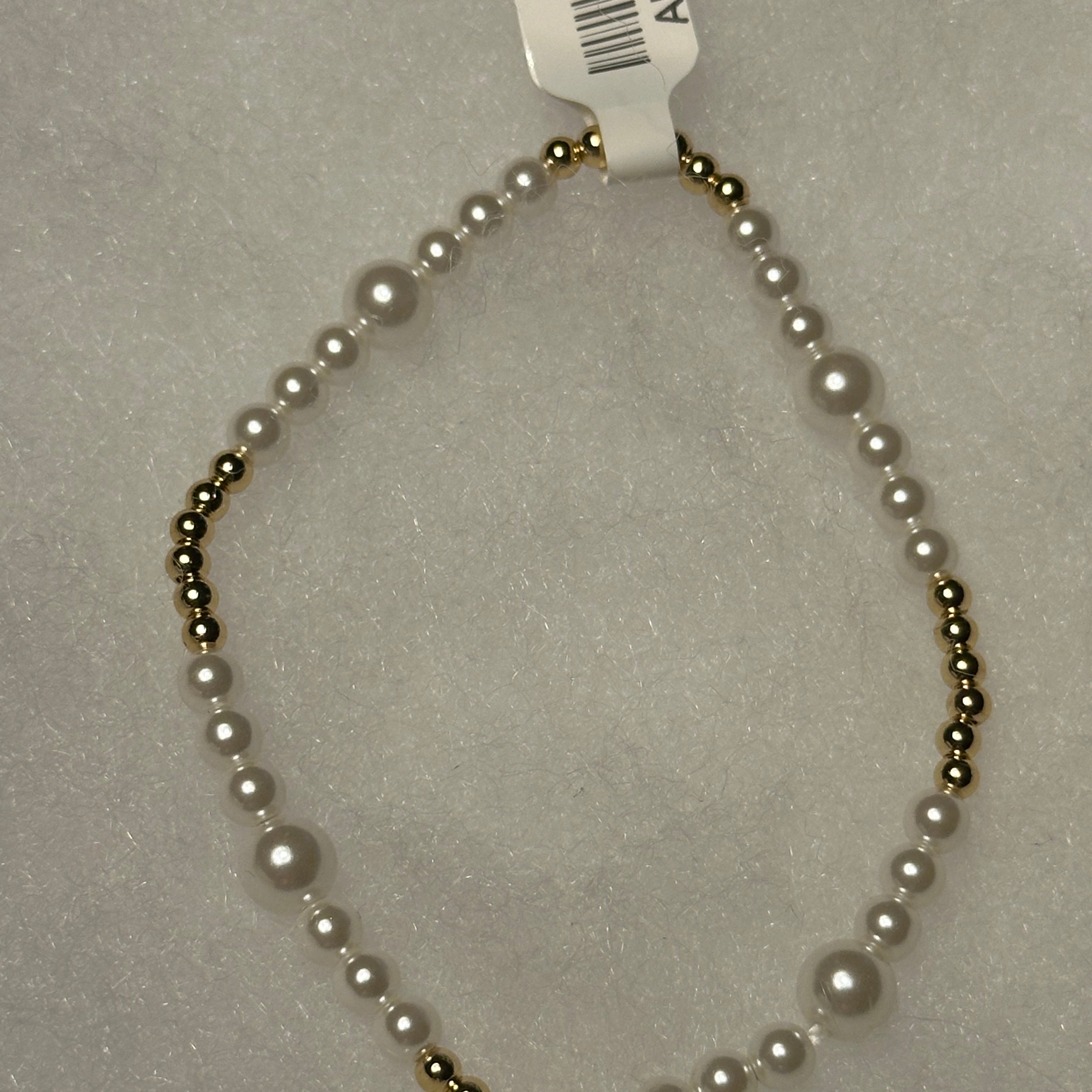A beaded necklace with white faux pearls and small metallic gold beads, shown on a light gray textured background with a white barcode tag attached.