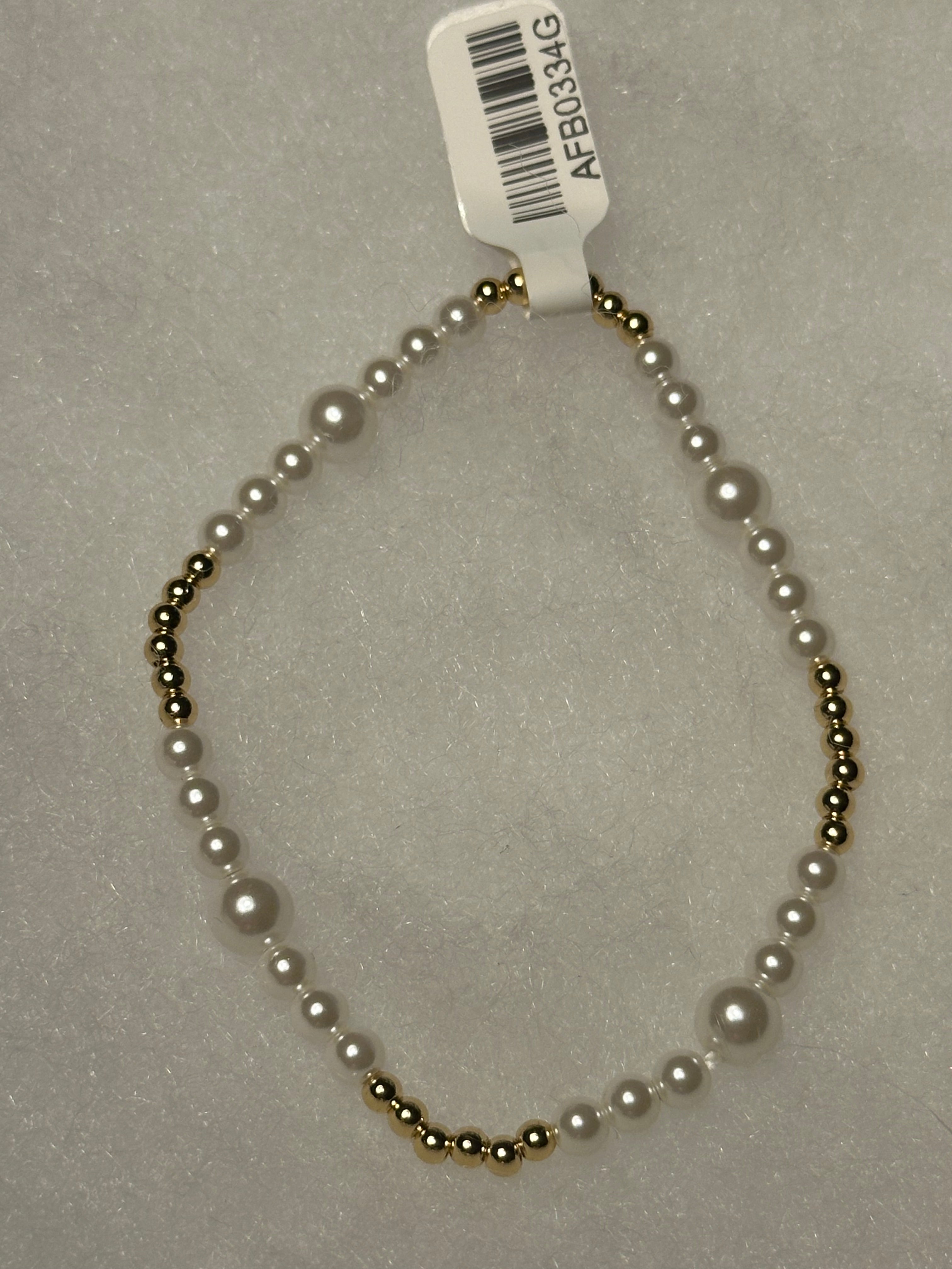 A beaded necklace with white faux pearls and small metallic gold beads, shown on a light gray textured background with a white barcode tag attached.