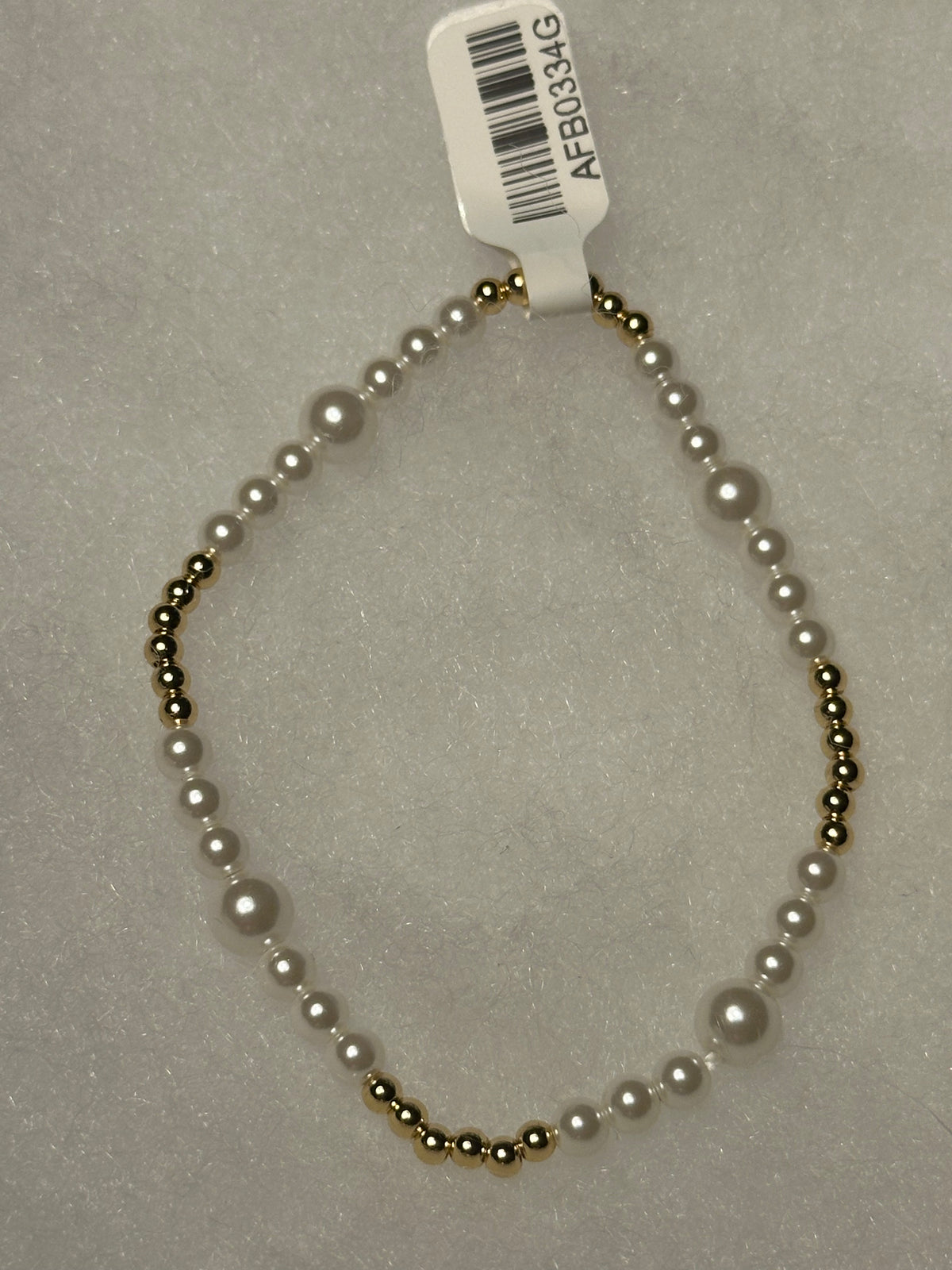 A beaded necklace with white faux pearls and small metallic gold beads, shown on a light gray textured background with a white barcode tag attached.