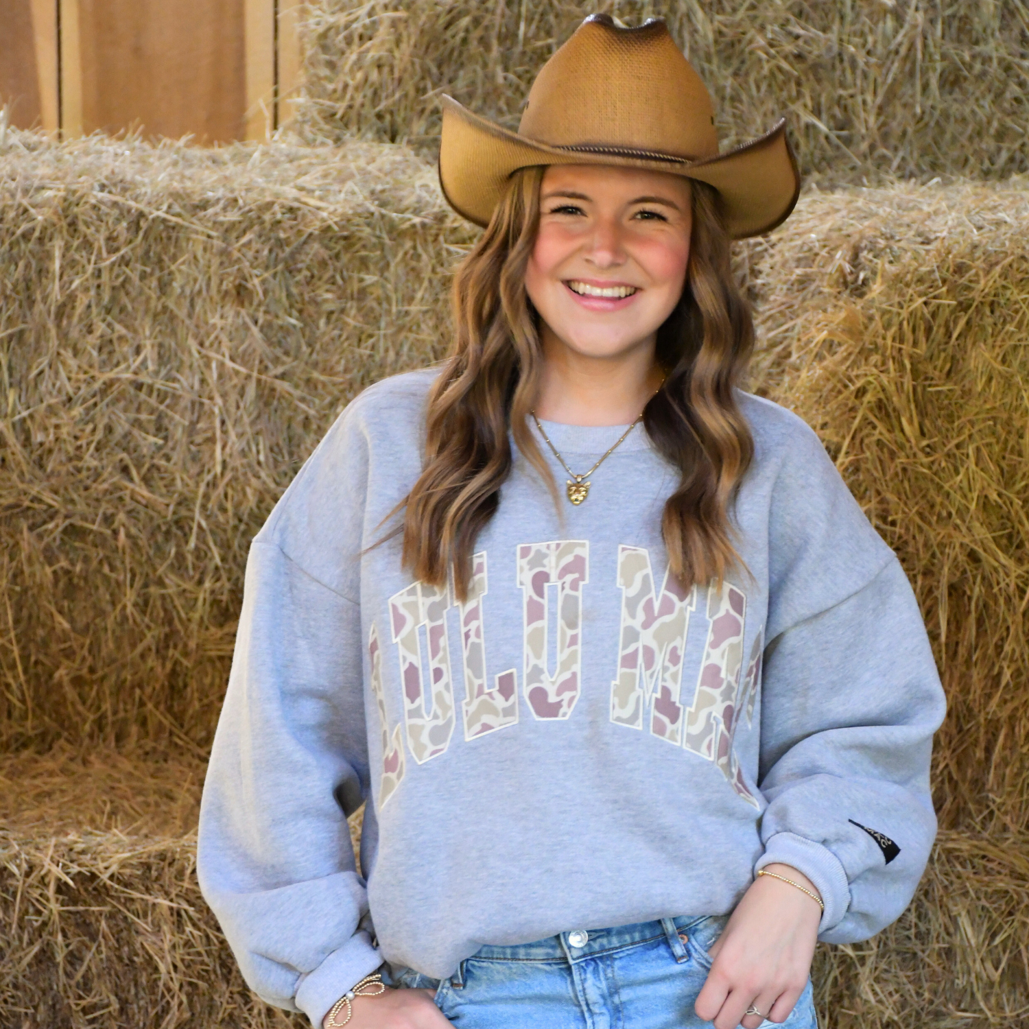 Person wearing a cowboy hat and patterned sweatshirt standing in front of hay bales.