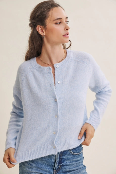 A woman wearing a light blue knit cardigan sweater with a button-up front, paired with blue jeans, against a plain beige background.