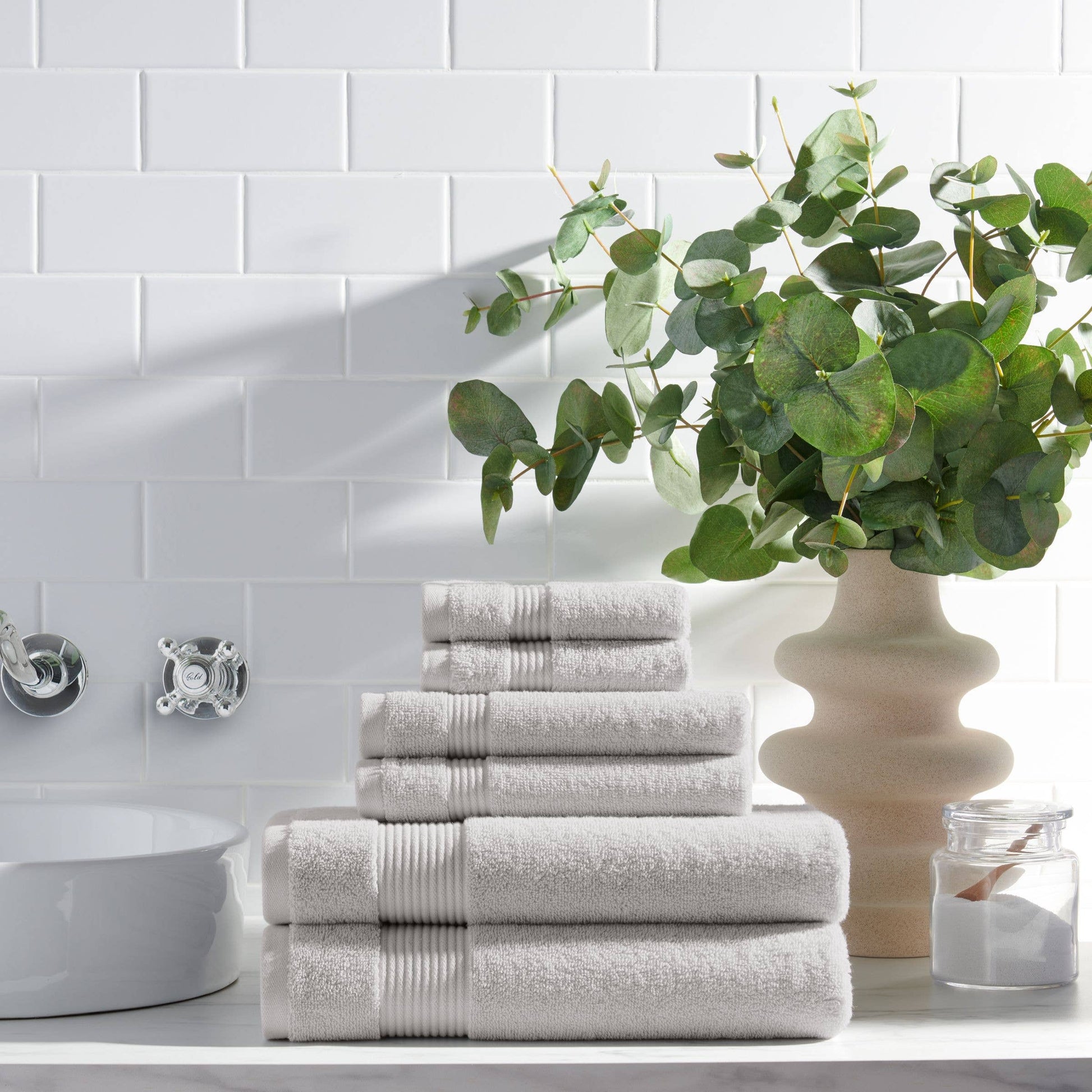 Stack of gray towels on a bathroom counter with a vase of greenery and white tiles in the background.