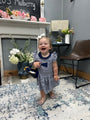 A toddler girl wearing a navy and white striped dress with ruffled sleeves and a bow in her hair, standing in a decorated room with a fireplace, chair, and rug.