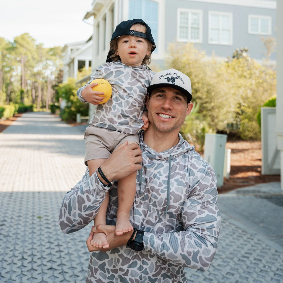 Man holding a child outdoors with a house and garden in the background