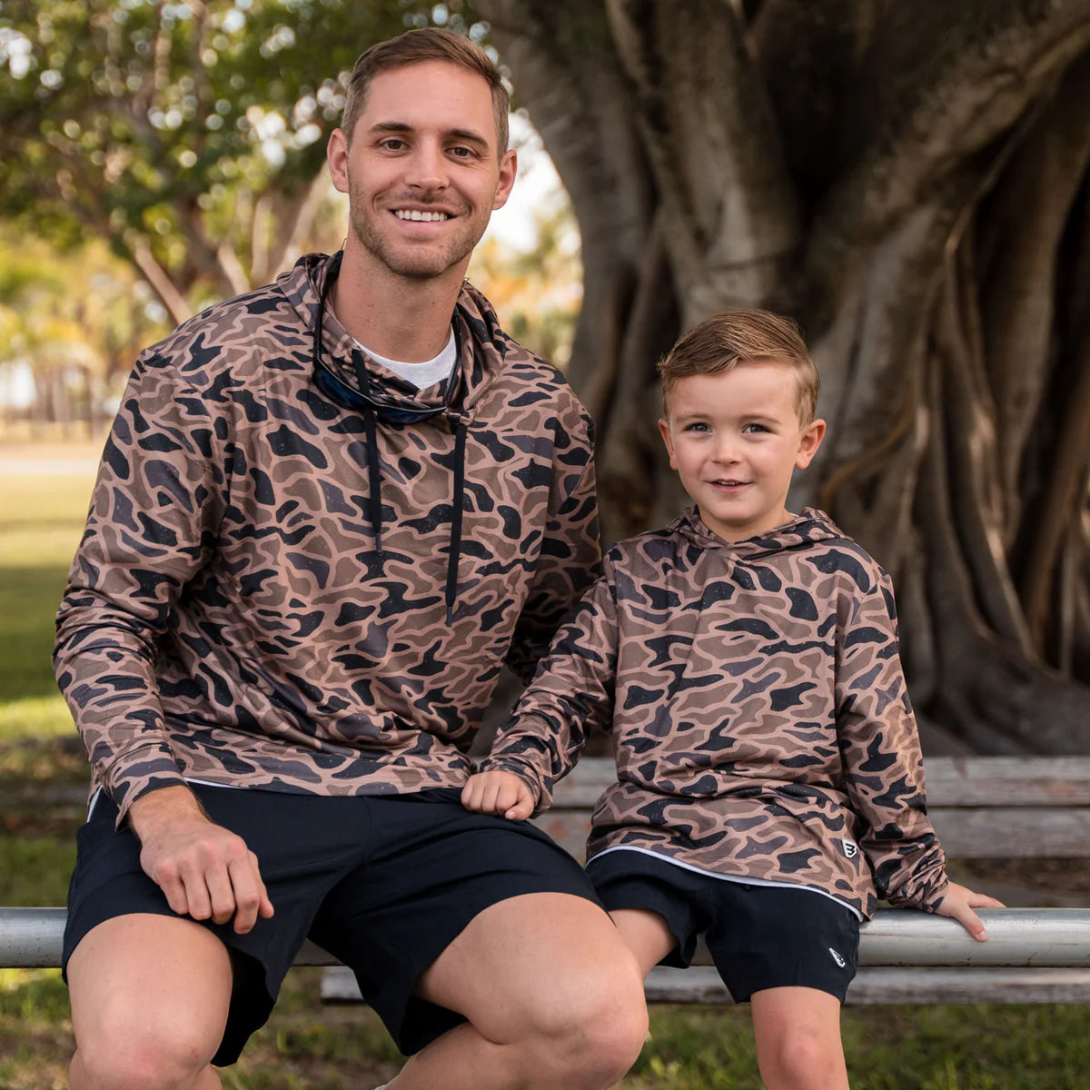 Man and child wearing matching camouflage hoodies sitting outdoors.